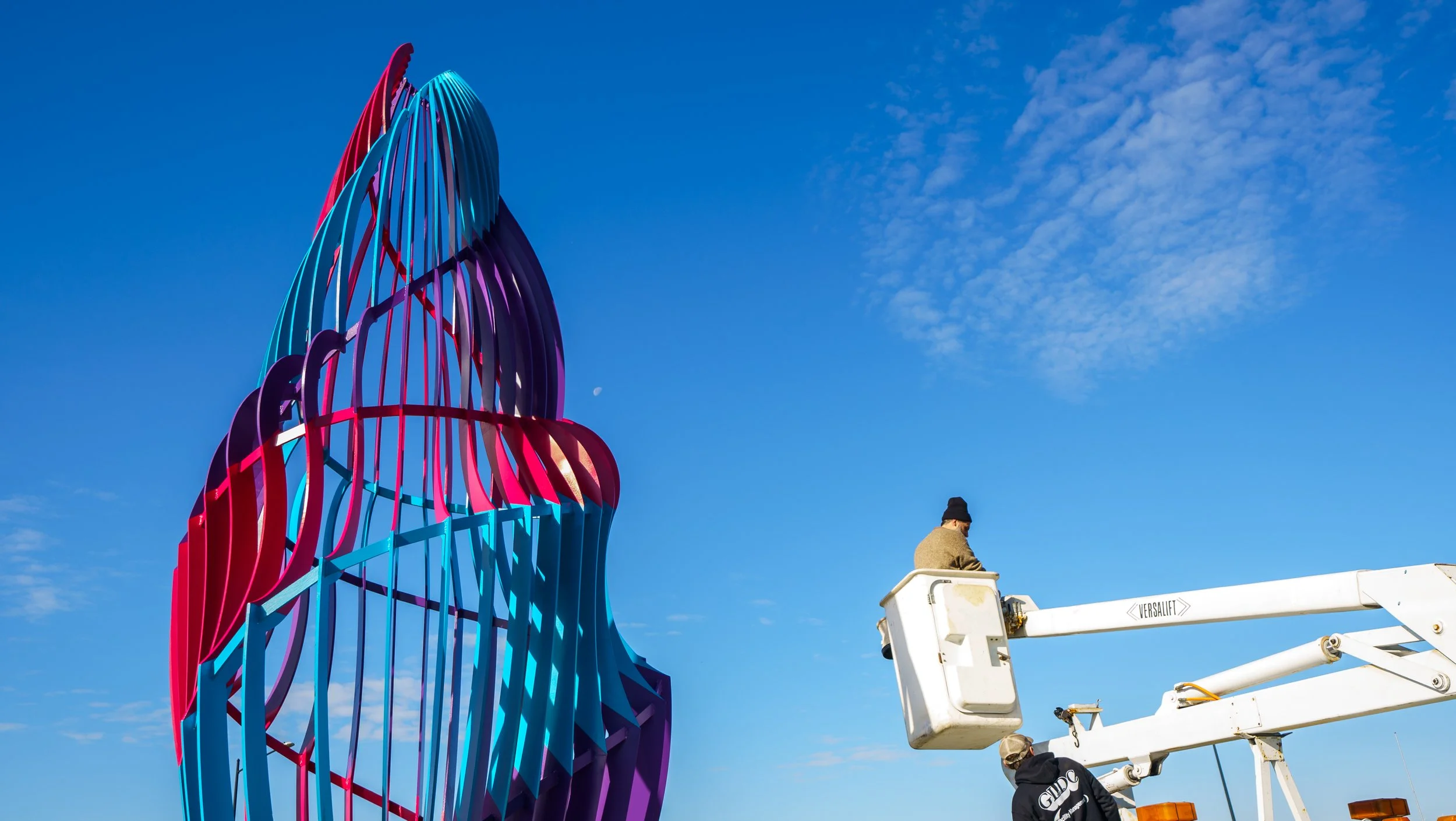 A person in a black jacket working on a large, colorful, abstract sculpture against a bright blue sky with scattered clouds.