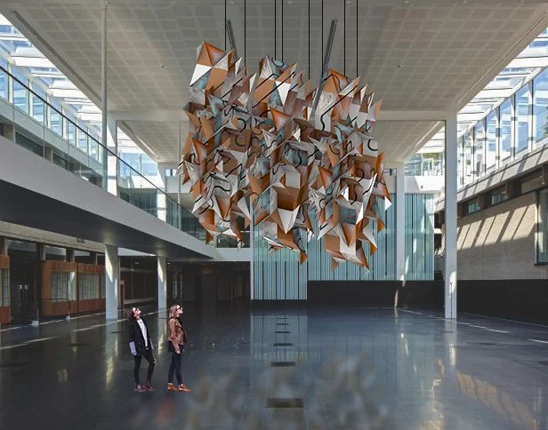 Modern interior with a large, abstract, geometric hanging sculpture in the center, featuring multiple interconnected shapes in wood, white, and blue. Two people are standing on the left side, looking at it.