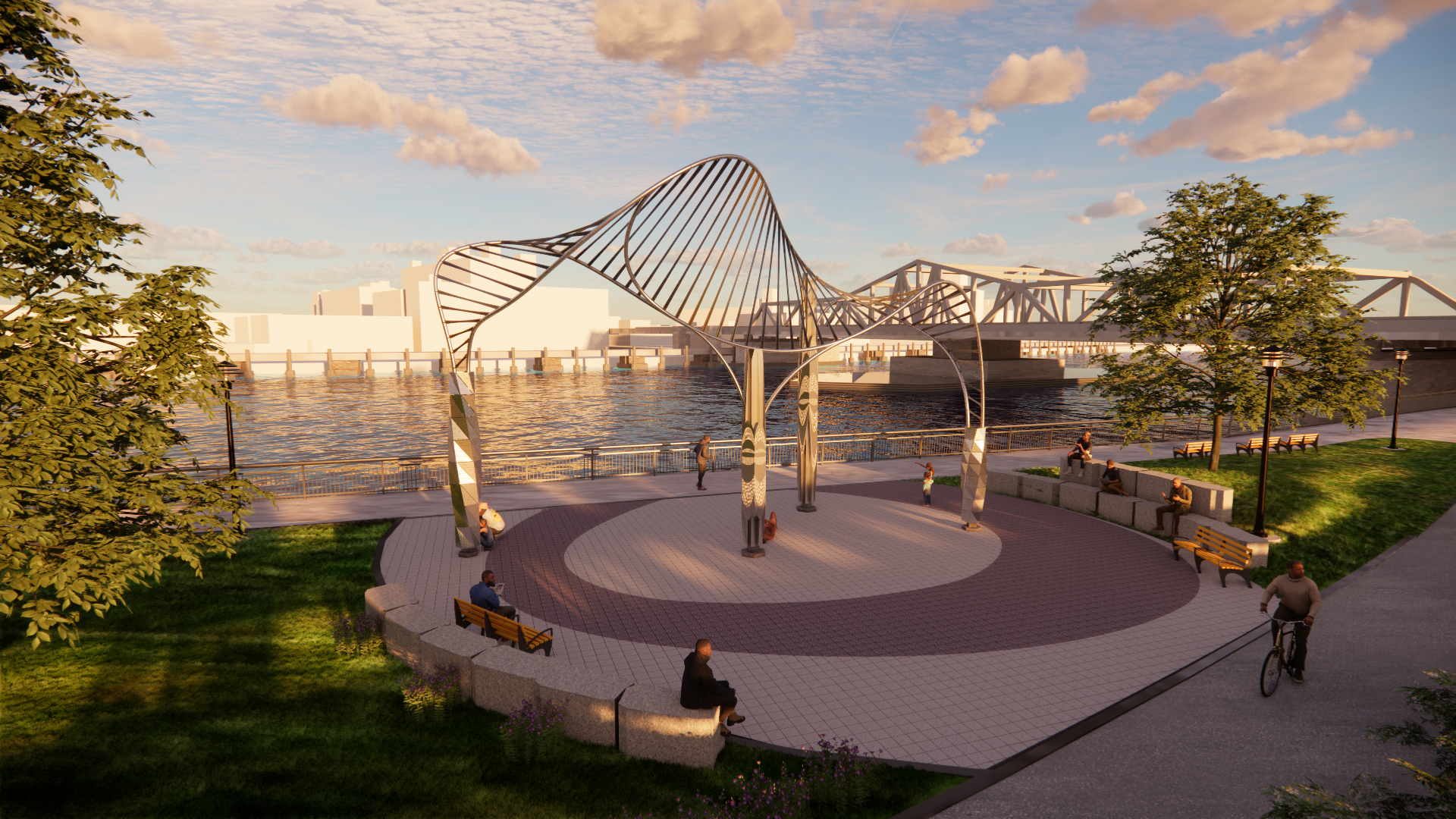 A park by the water with a modern sculpture, benches, and trees, with a bridge and city buildings in the background during sunset.