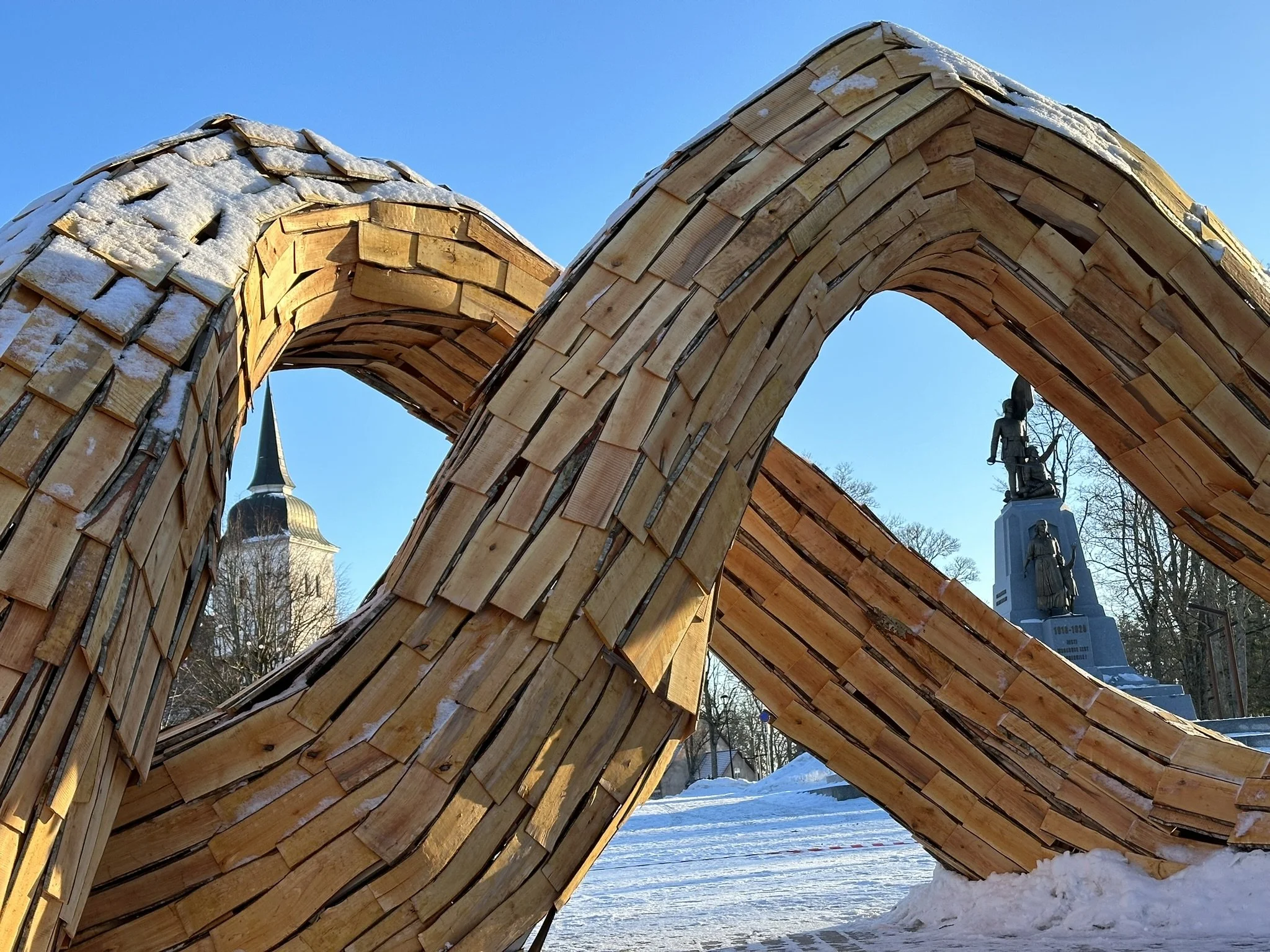 Close-up of a large wooden sculpture shaped like an abstract star or snowflake with snow on top, set outdoors under a clear blue sky.