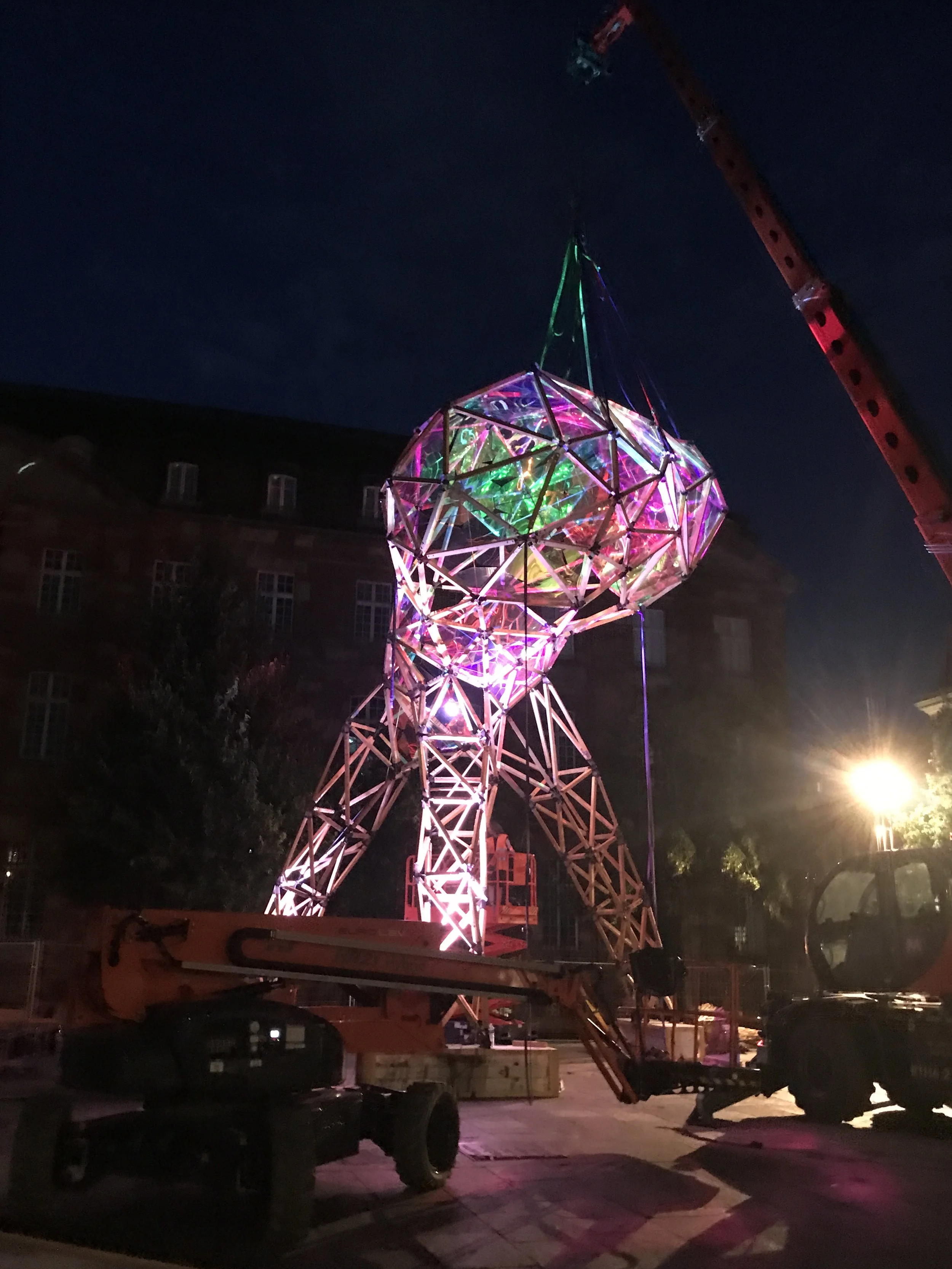 A large geometric structure illuminated with colorful lights is being assembled outdoors at night. A crane is lifting the structure, and there are several vehicles and equipment around.