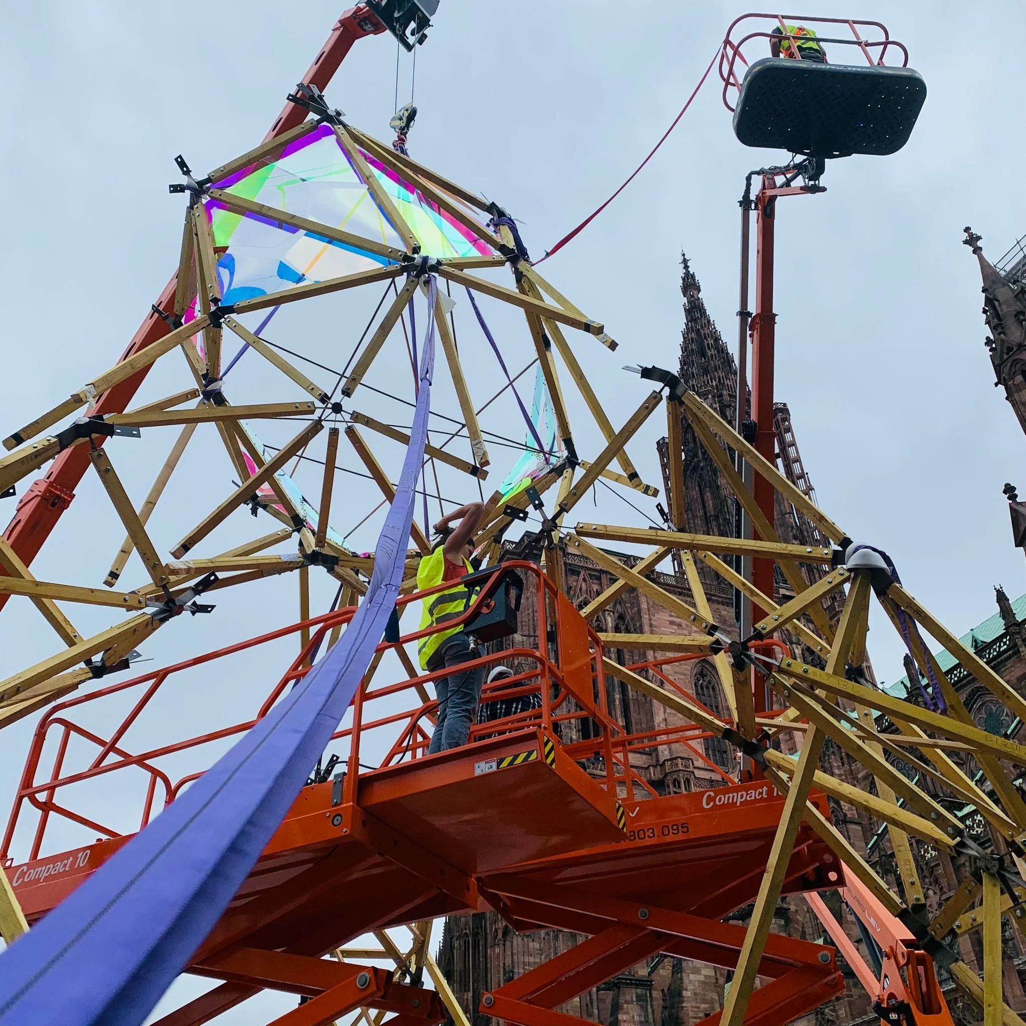 Construction workers on an orange scaffolding structure building a large, colorful installation in front of a historic brick building with pointed spires.