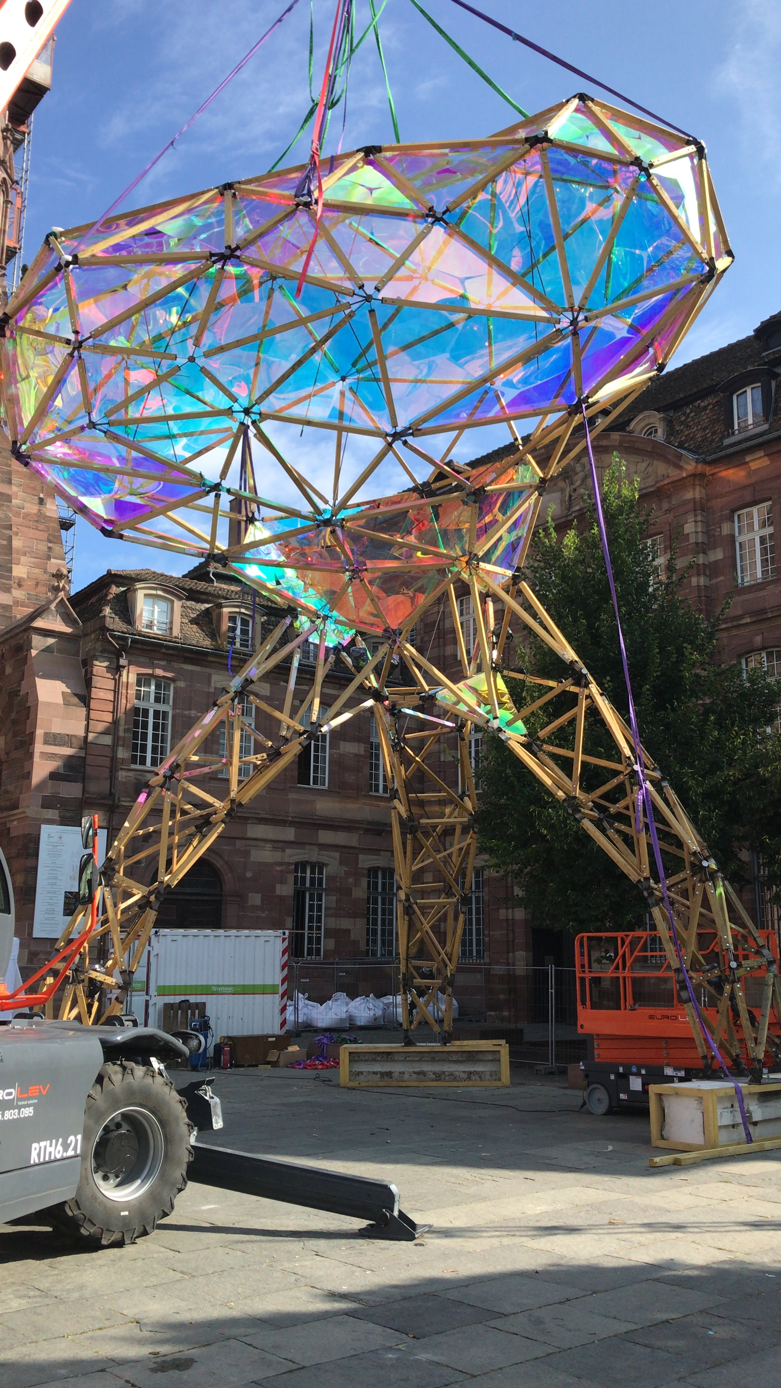 Construction site with a large sculptural structure made of wooden beams and iridescent panels, set against historic brick buildings and a partly cloudy sky.