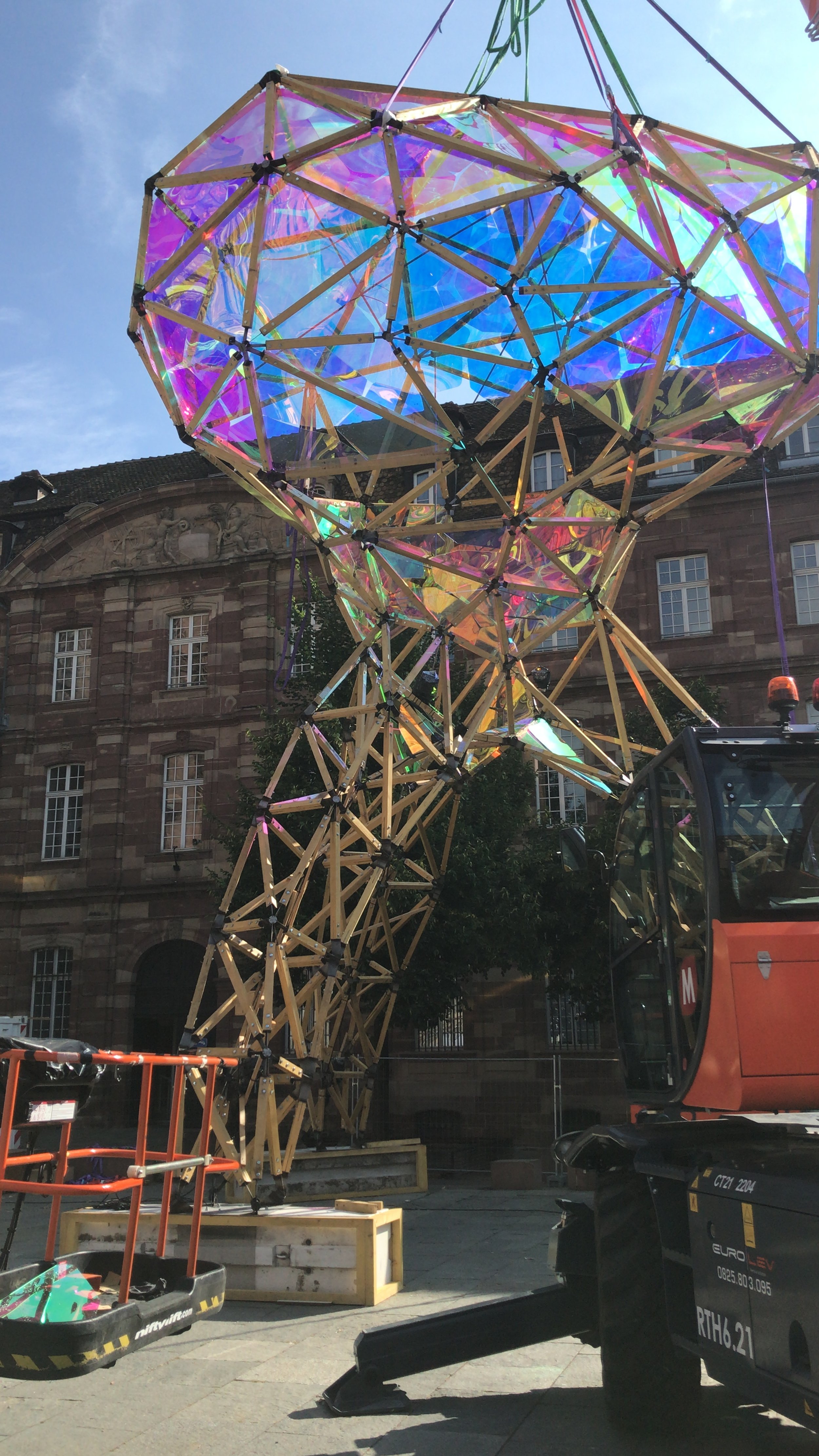 A large geometric installation made of wooden sticks and iridescent panels suspended in the air, with parts of the structure visible on the ground. The installation is outdoors in front of a historic brick building, with equipment and a construction 