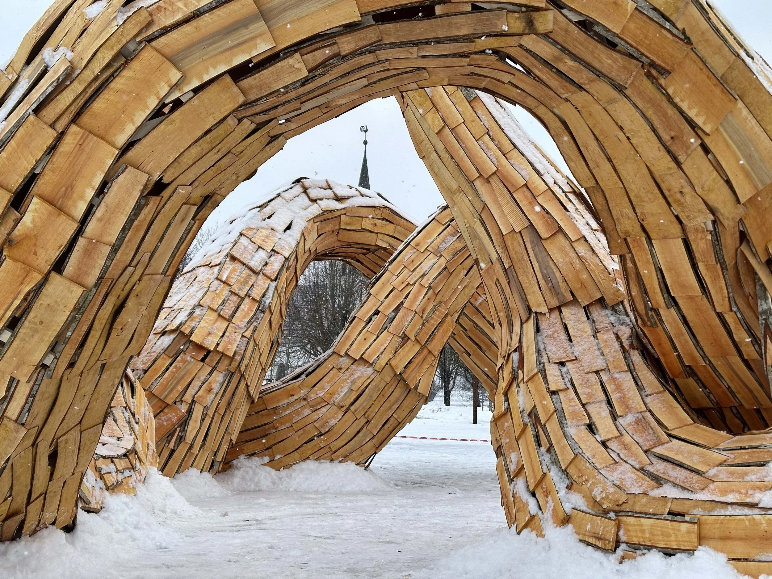 A large wooden sculpture with intertwined, curved wooden planks covered in snow, located outdoors in a snowy landscape with a church steeple visible through the opening of the sculpture.