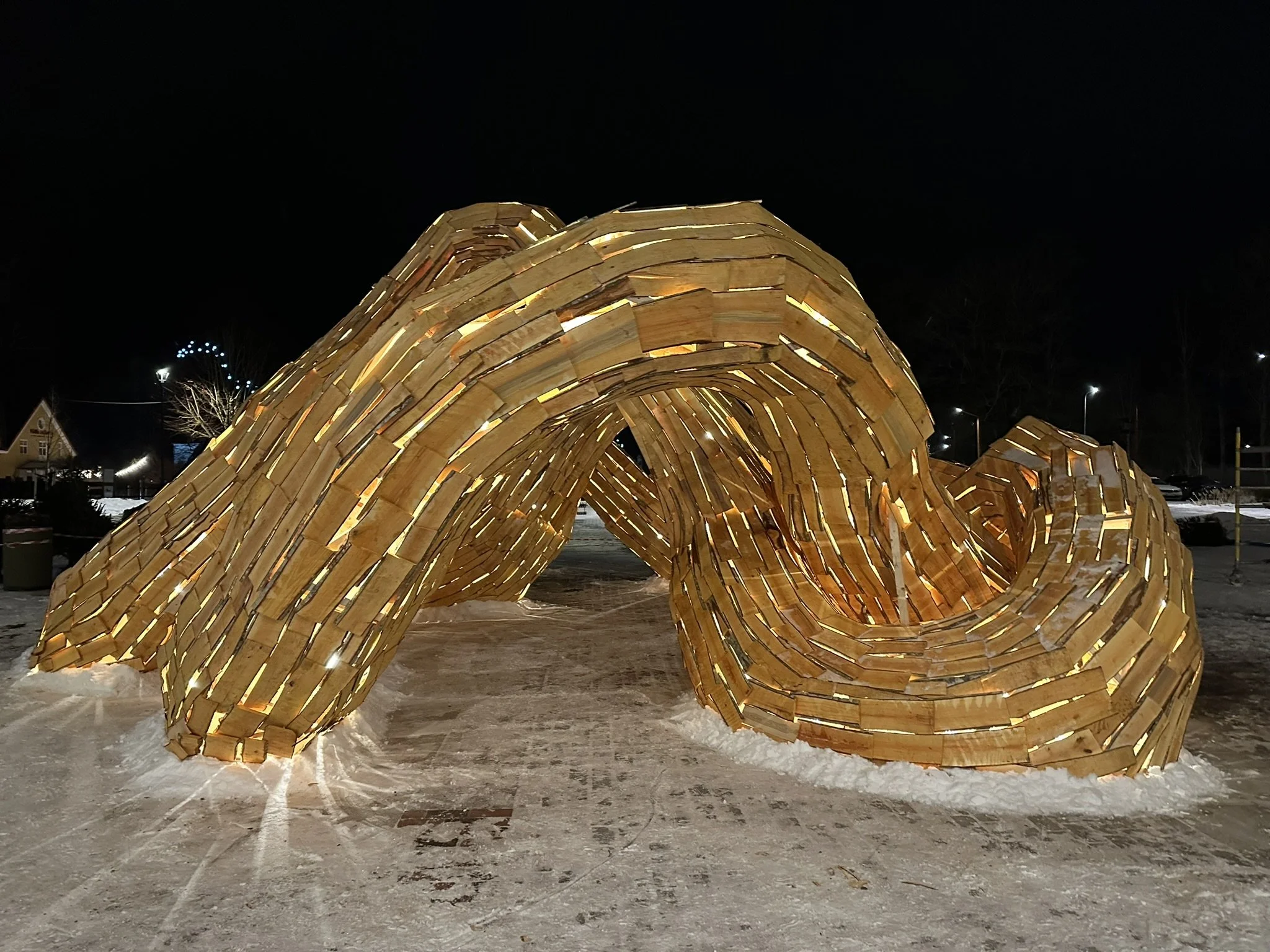 Night scene of a large, illuminated, wooden sculpture resembling a curled or coiled shape outdoors with snow on the ground.