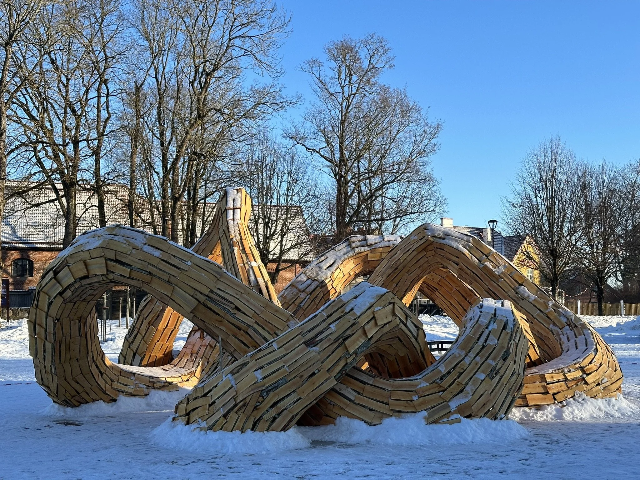 Wooden sculpture of linked circles and curves outdoors in a snowy park with leafless trees and buildings in the background on a clear, sunny winter day.