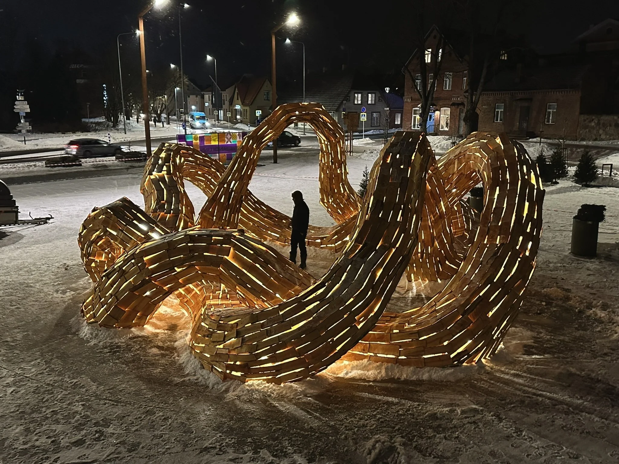 Nighttime scene in a snowy town square with illuminated wooden sculpture resembling intertwined chairs and pumpkins, and a person standing nearby.