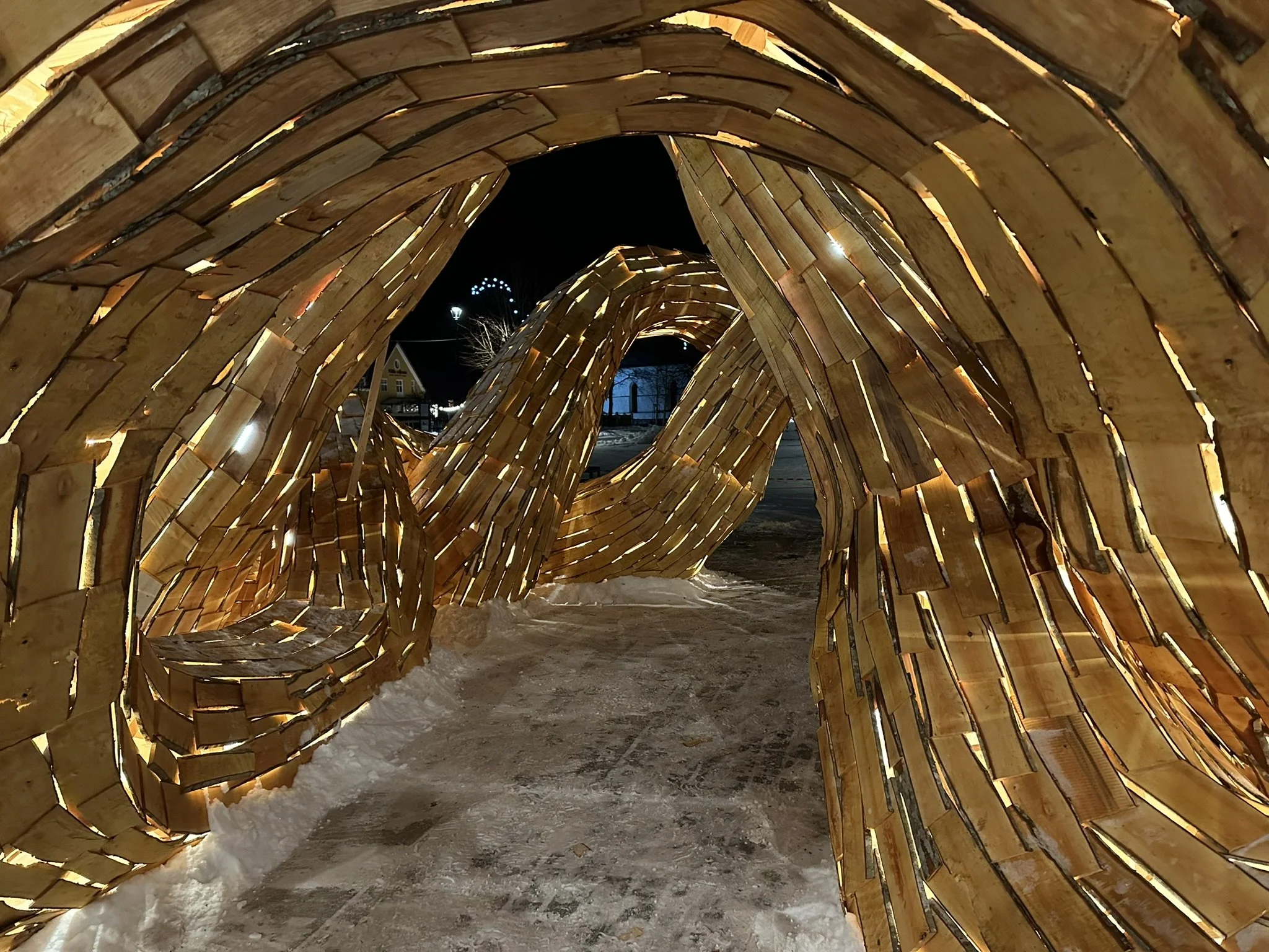 Wooden tunnel sculpture at night, illuminated from within, with snow on the ground and a house and trees visible in the background.