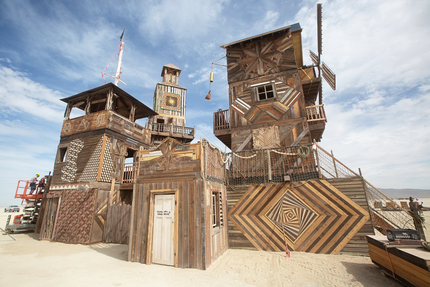 A large, quirky wooden structure built on a sandy area, possibly a park or art installation, with multiple levels, towers, and geometric patterns on the walls. The sky is partly cloudy with a few people visible on the left and right sides.