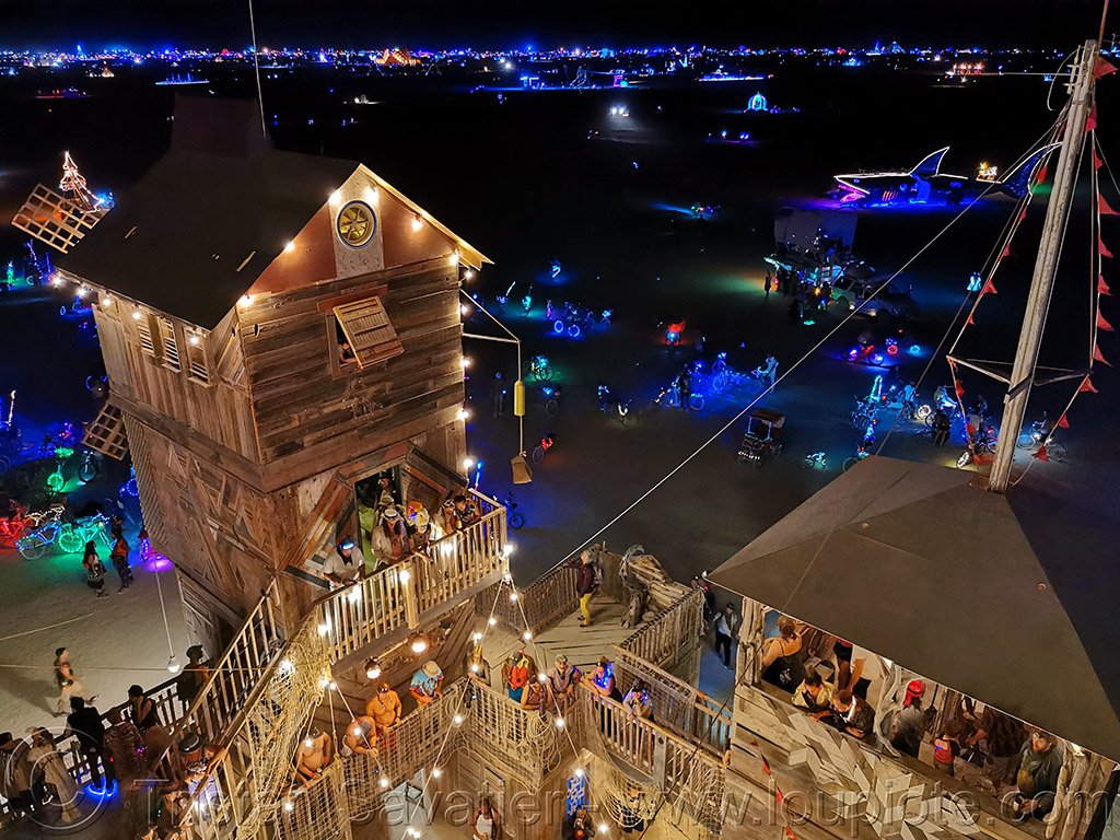 Night scene of a festival with a wooden tower, colorful lights, people, and decorated bikes.