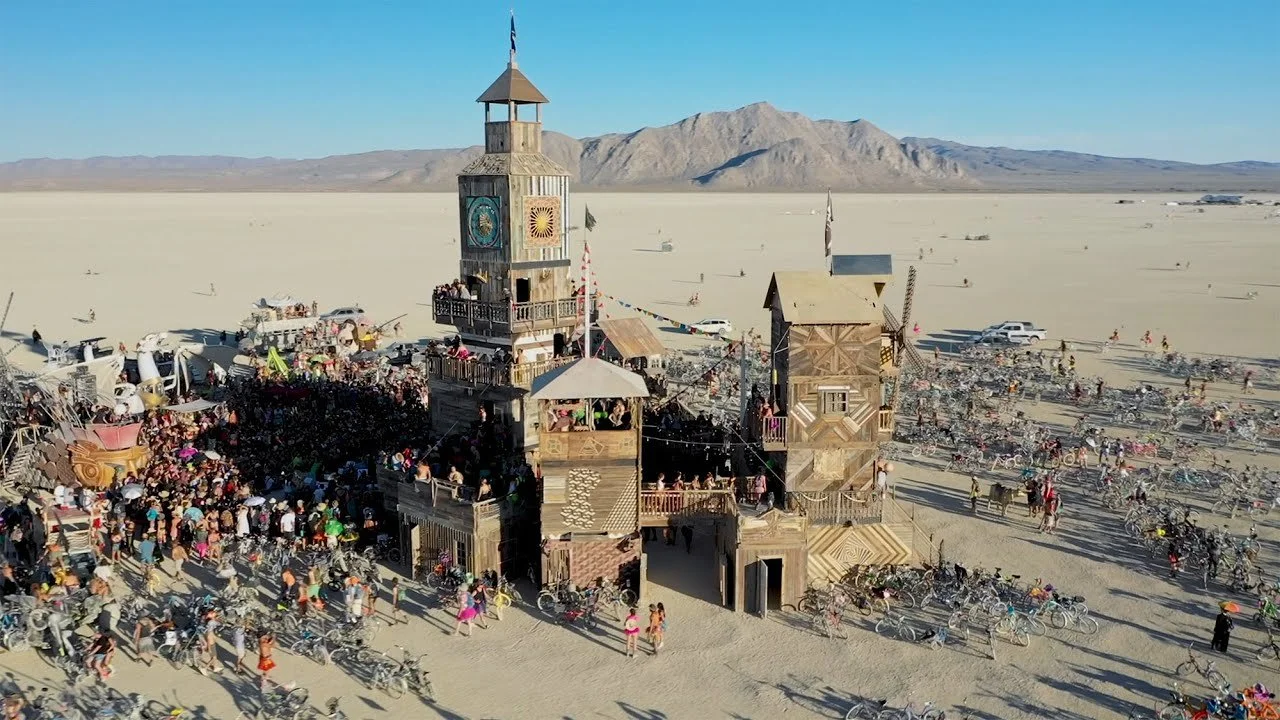 A large wooden structure with towers and balconies in a desert, surrounded by many people, bicycles, and tents, with mountains in the background.
