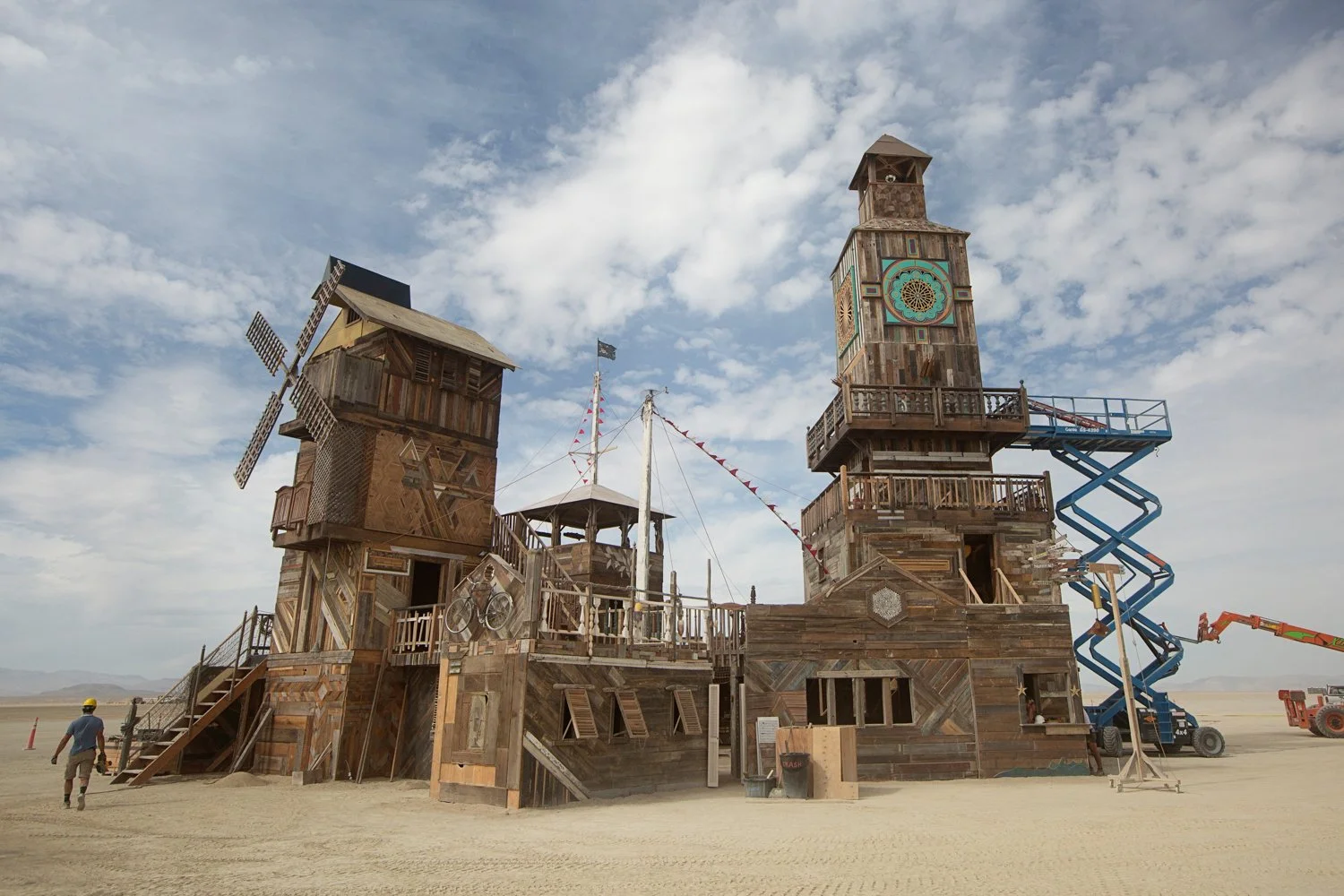 A large art installation resembling a wooden castle with multiple towers, set in a desert. The castle features a windmill on one tower, a clock on another, and a slide. A person with a yellow helmet and a person with a hat are nearby, with constructi