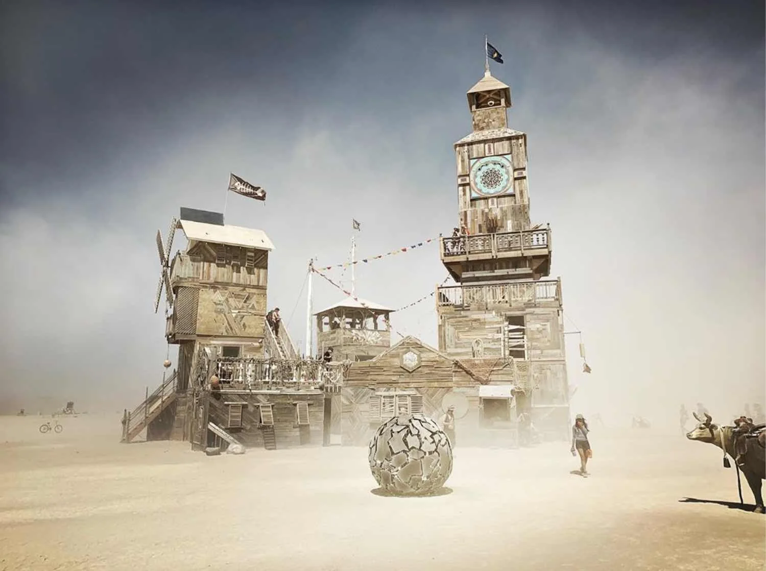 A large wooden structure resembling a town or fort on a dusty, barren landscape with a large spherical object with cracks in the foreground. There are people walking around, some on stilts, and a couple of animals, including a cow. The sky is overcas