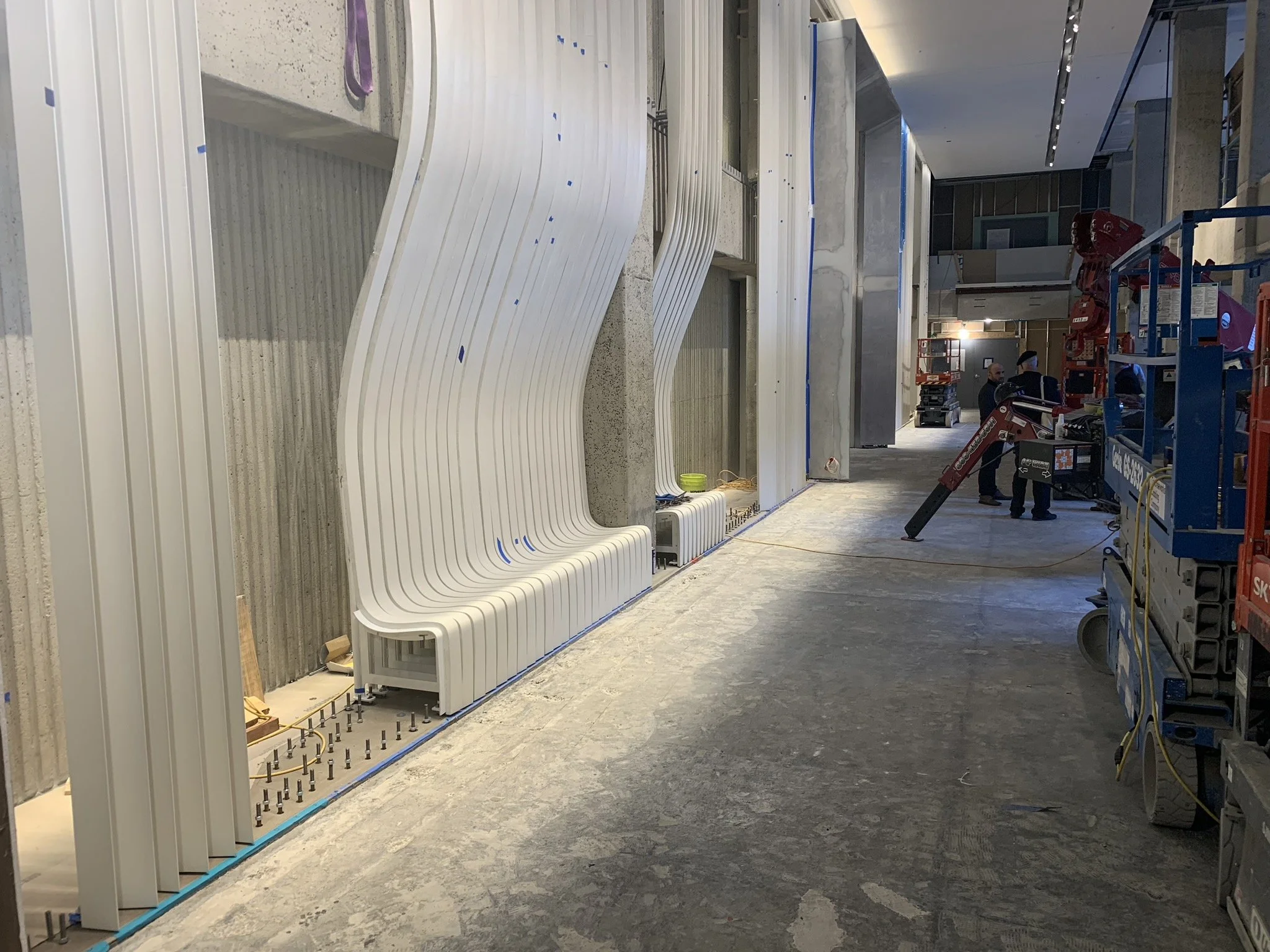 Inside a building under construction, white curved architectural panels are being installed on a wall. Construction workers and equipment are visible in the background.
