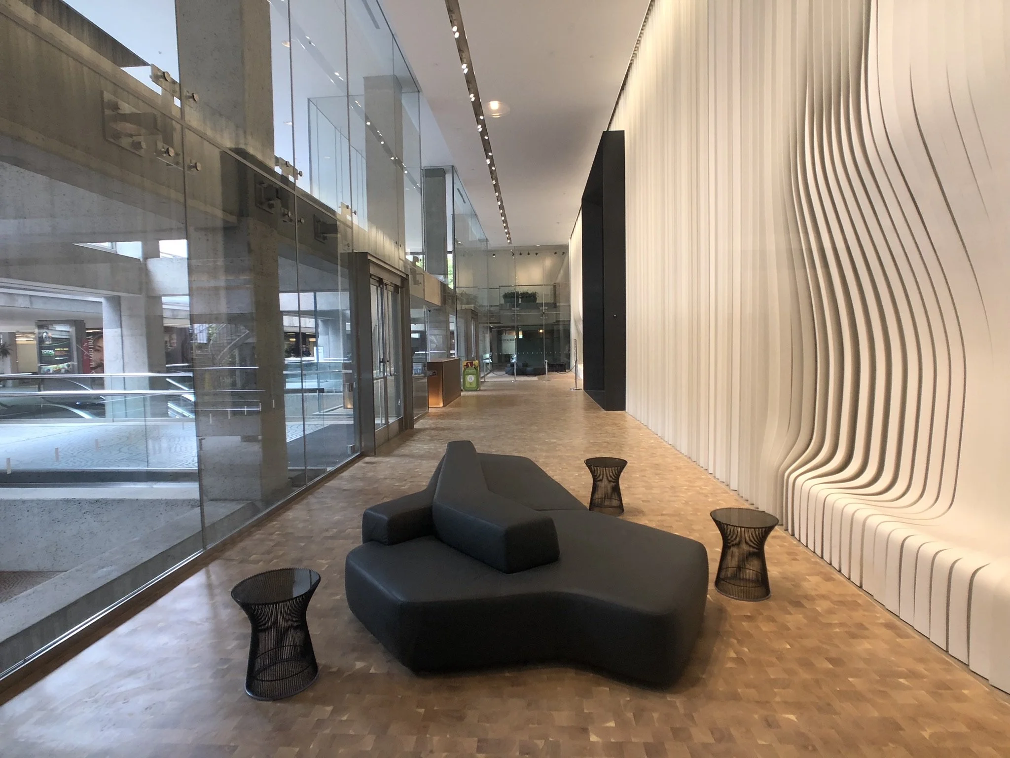 Modern interior space with a large glass wall, a curved textured white wall, black seating with round side tables, and a wooden floor.