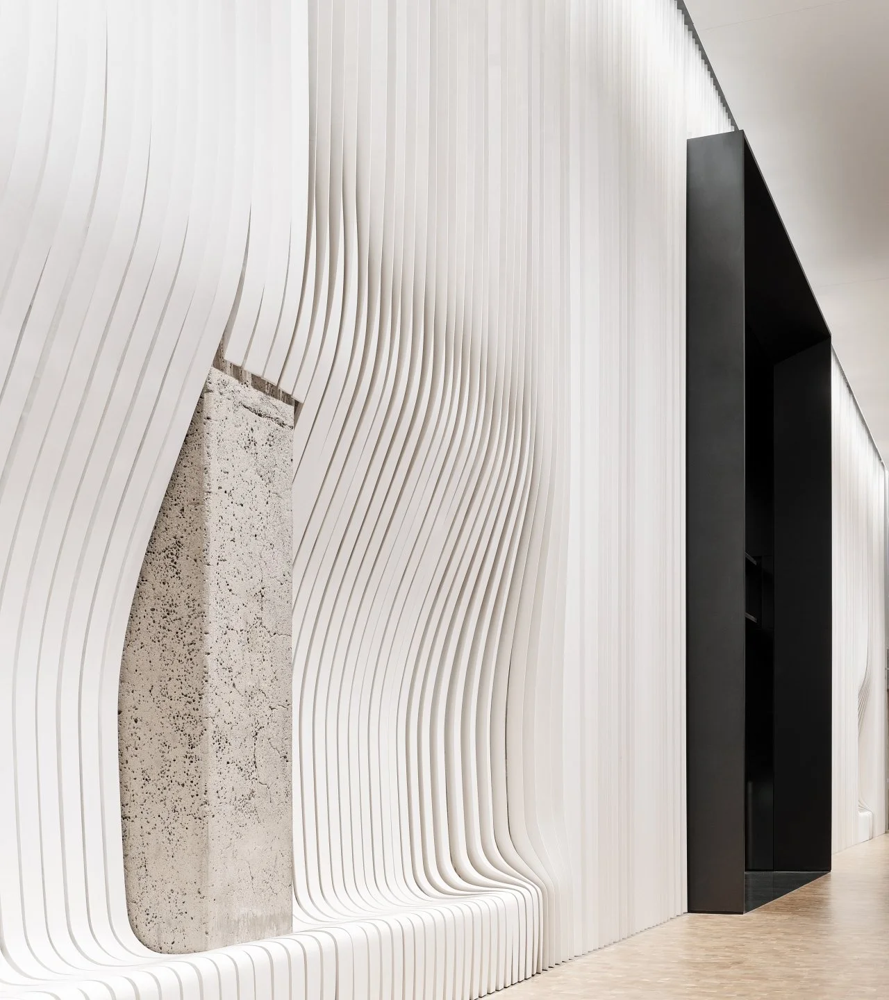 Modern interior wall with wavy white panels, a textured concrete pillar, and a black rectangular doorway.