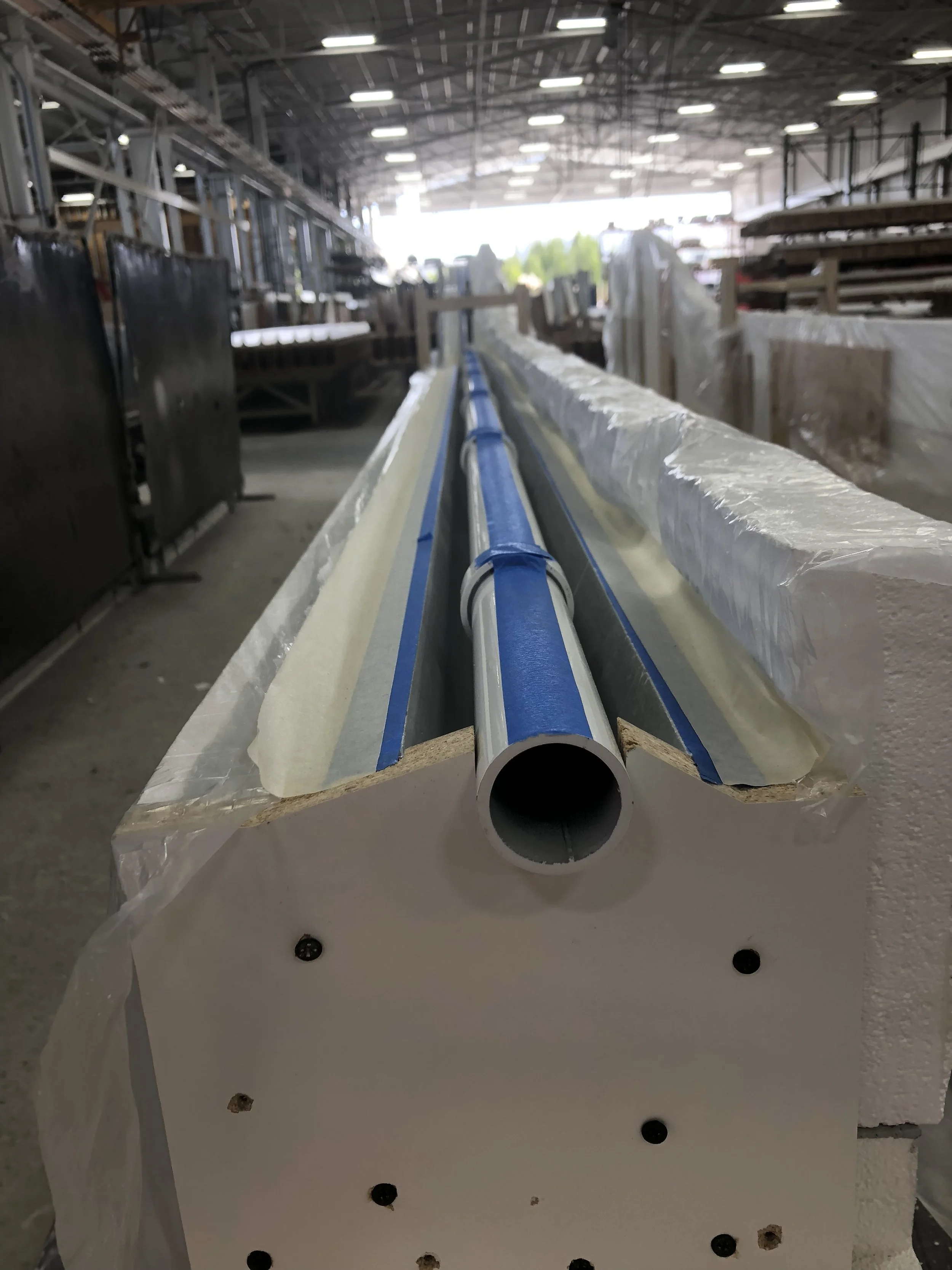 A long metal pipe is positioned on a white surface, with a plastic coating on it. The pipe is placed in front of a conveyor system inside a large industrial warehouse with a high ceiling and metal framework. The warehouse has various stacks of materi