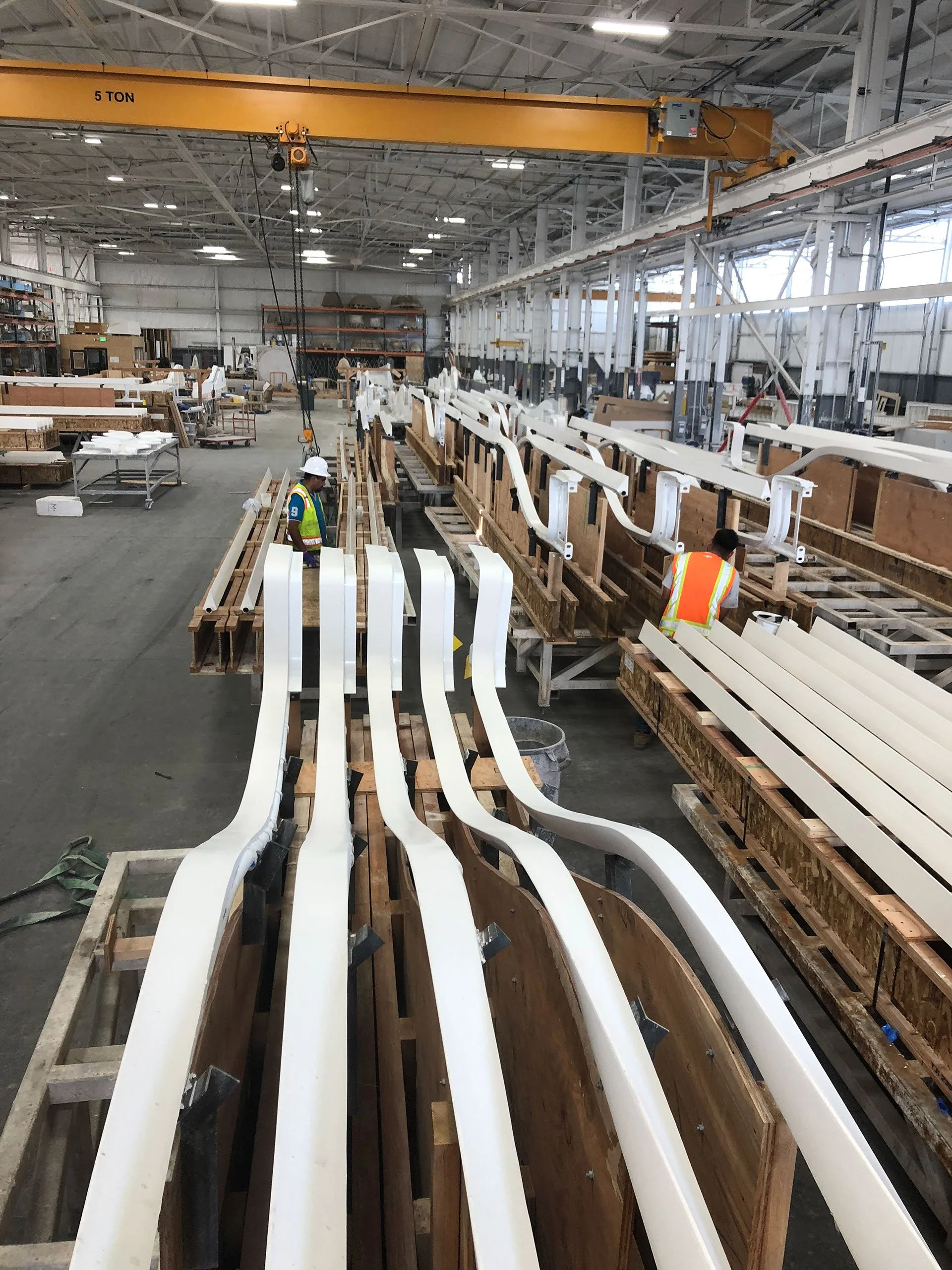 Inside a manufacturing facility with large wooden molds and white curved parts being assembled, with two workers wearing safety vests and helmets, and a yellow overhead crane labeled '5 TON'.