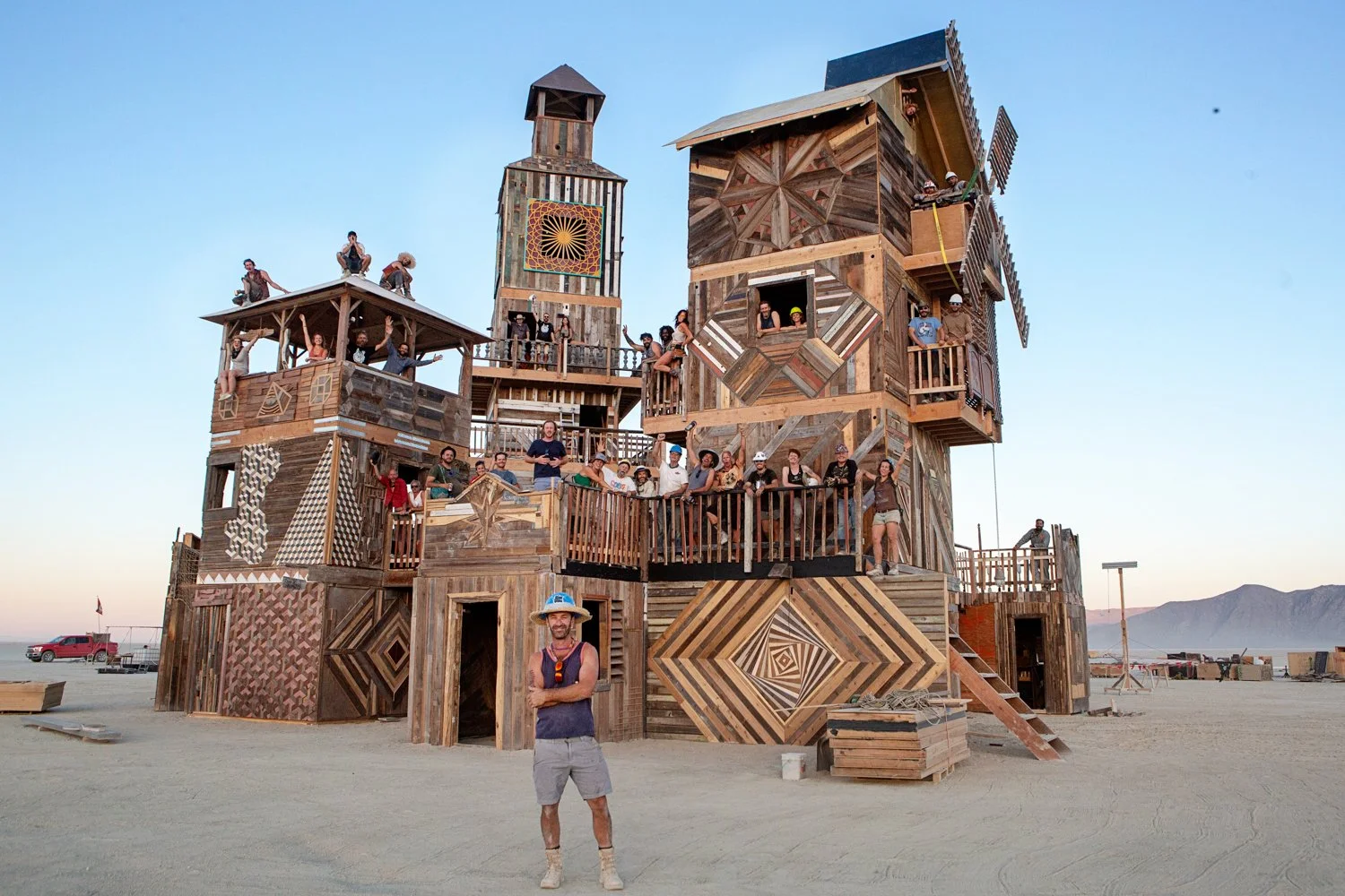 Large multi-story wooden art installation with geometric patterns, situated in a desert with mountains in the background. Many people are standing, sitting, or climbing on the structure, and a man with a hat and sunglasses is standing in front of it.