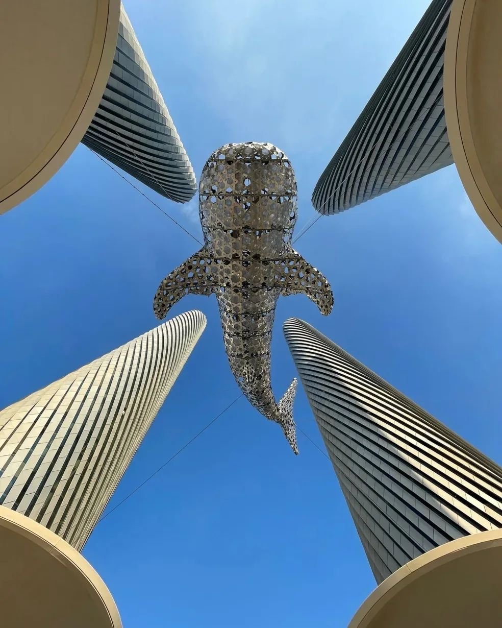 A large, metallic shark sculpture suspended between four tall, modern skyscrapers with curved facades against a clear blue sky.