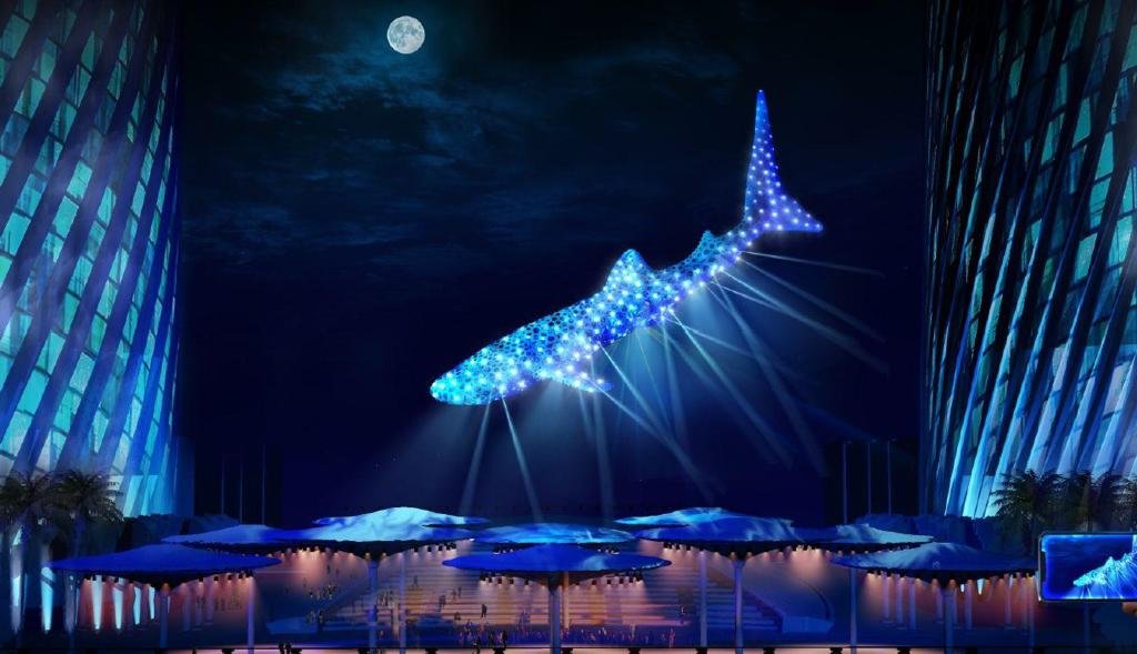 A large illuminated whale-shaped sculpture glowing blue, suspended in the night sky with the moon visible above. The scene is set at a modern outdoor structure with blue-lit walls and some trees in the background.