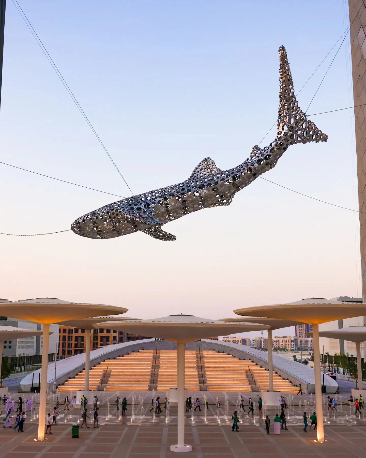 Large whale-shaped sculpture suspended in the air outdoors, with people walking below and modern structures in the background.