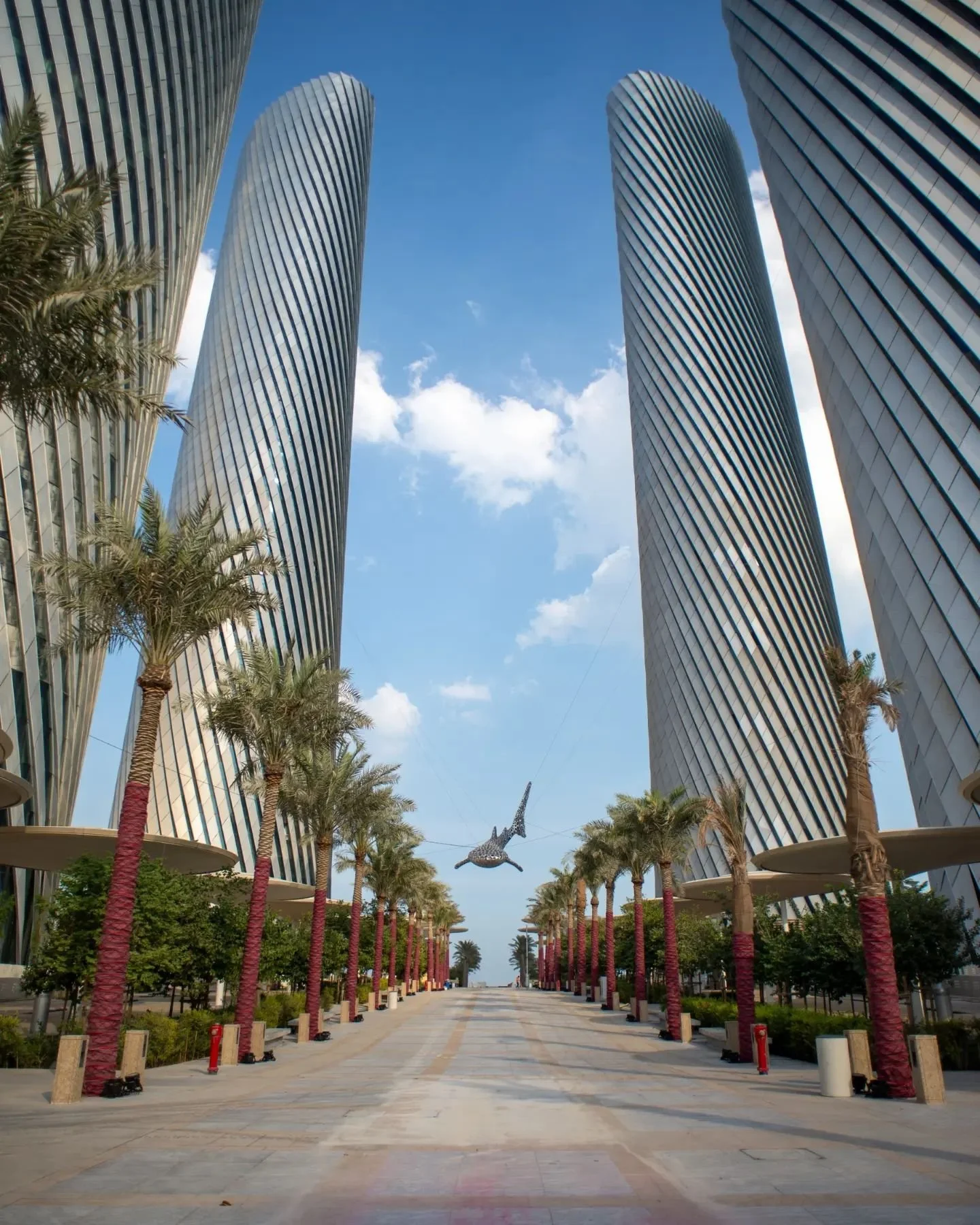 A walkway lined with palm trees between modern skyscrapers under a blue sky with clouds, and a flying sculpture resembling a flying fish.
