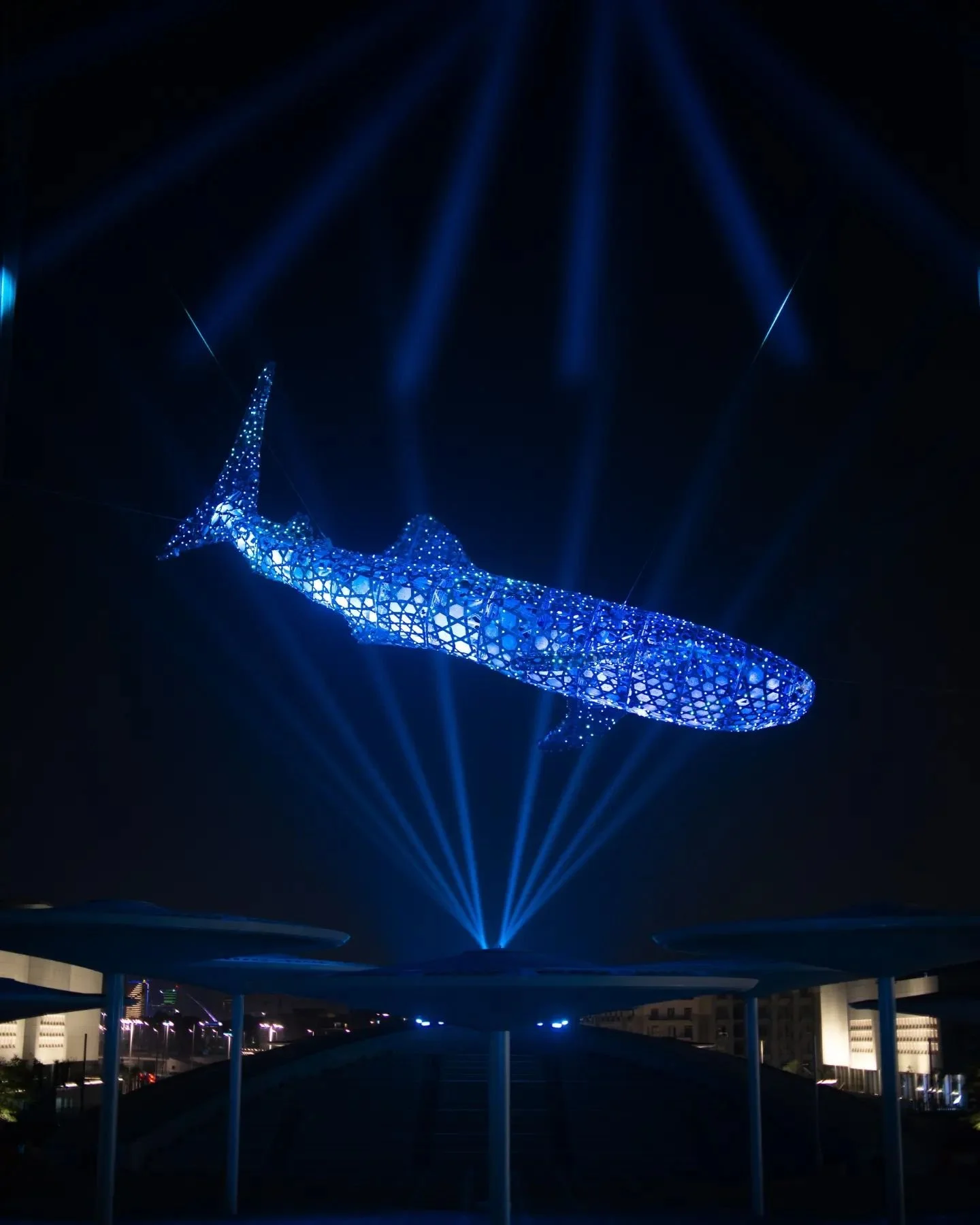 A large illuminated sculpture of a whale with blue lights, suspended from the ceiling at night, surrounded by blue spotlights.