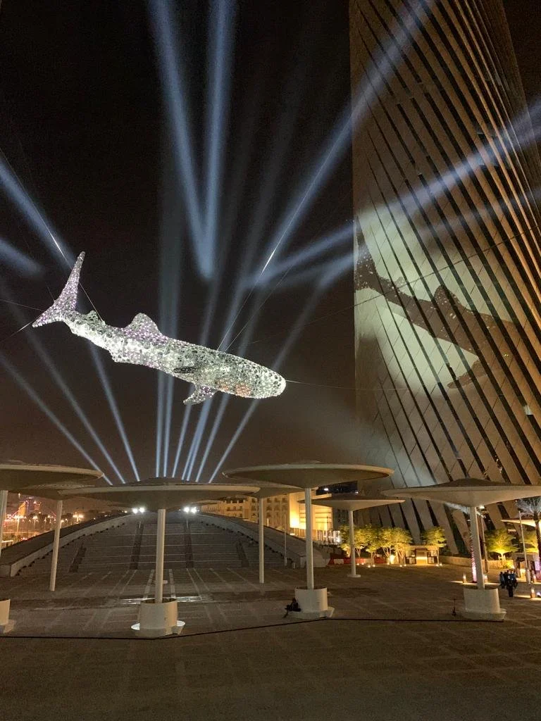 Nighttime scene with a large illuminated whale sculpture suspended in the air, with beams of light projecting upwards against a dark sky. The whale appears to be made of sparkling material, and there is a modern building with slanted design and verti