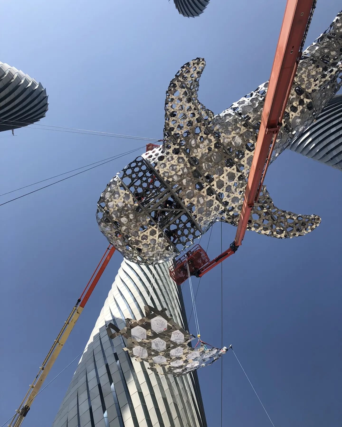 Construction workers installing a large, modern architectural sculpture on top of a tall building against a blue sky.