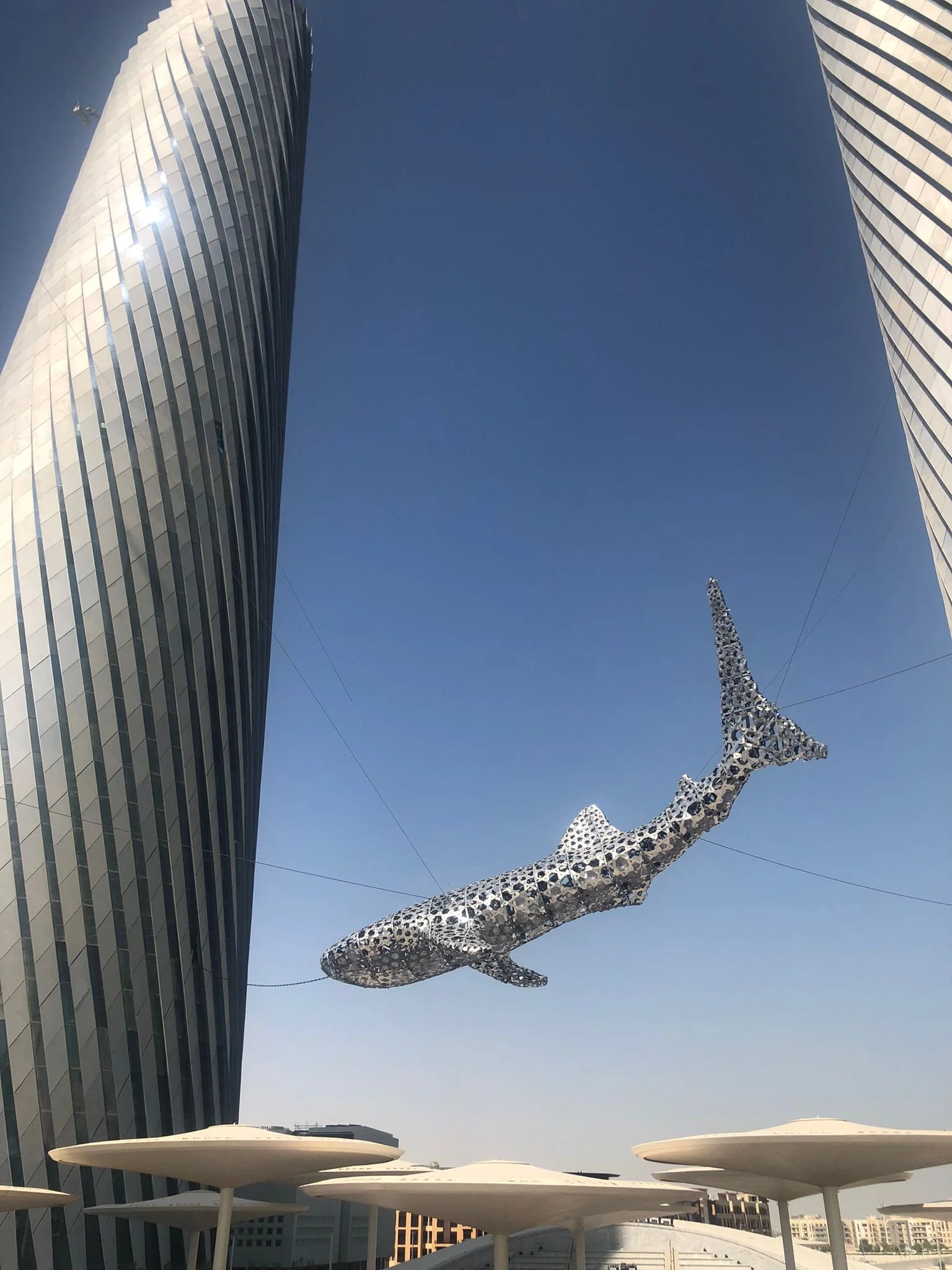 Tall modern skyscrapers with reflective glass facades, clear blue sky, and a metallic whale sculpture suspended in mid-air, with white canopy structures at the base.
