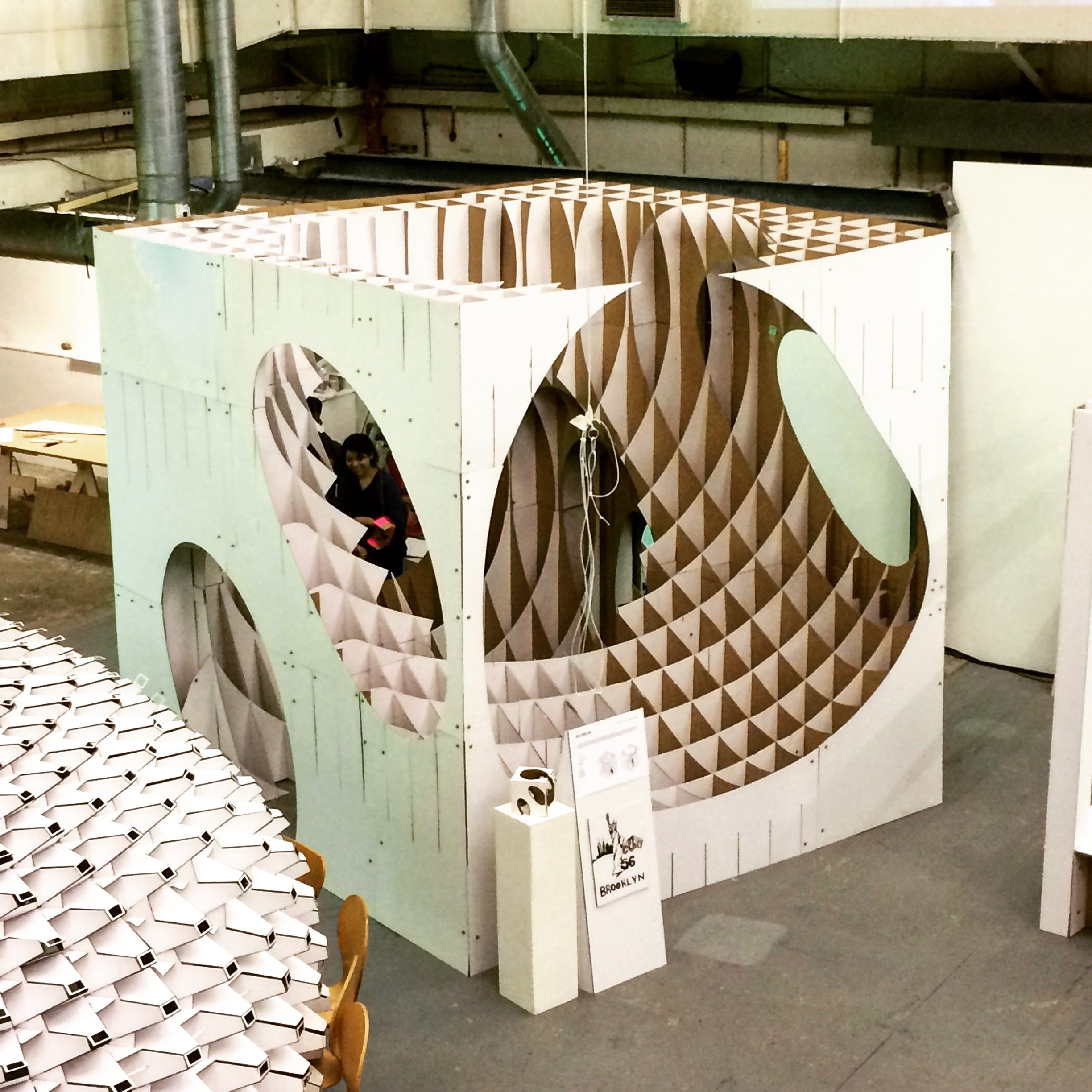 Interior of a space with white hexagonal tables and a large decorative structure made of wood in the shape of a face or animal head, with an opening for an eye and geometric patterns. A person is partially visible inside the structure.