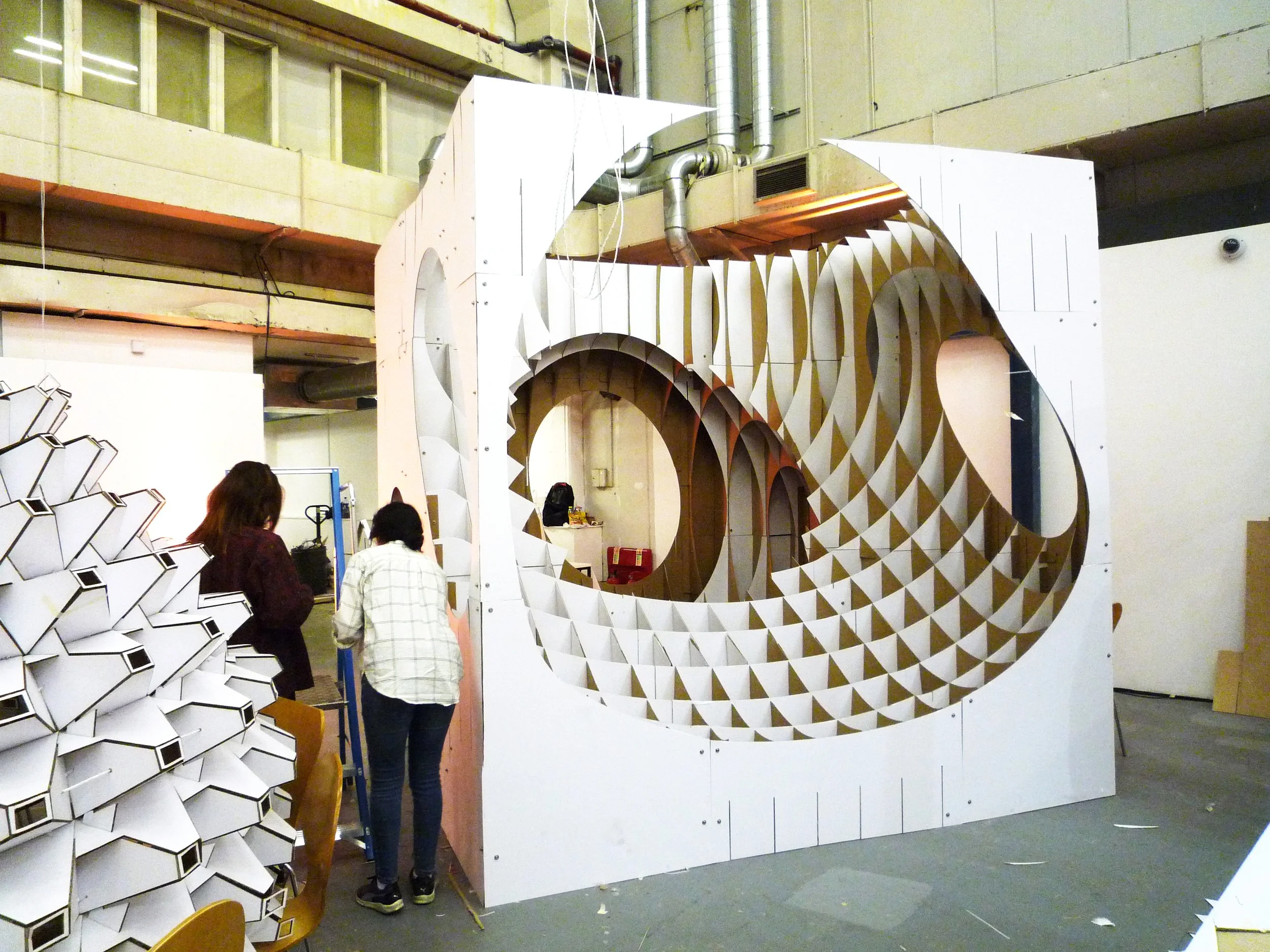 People standing near a large abstract geometric art installation made of white and brown panels with large cutouts, inside an industrial space with pipes and concrete walls.