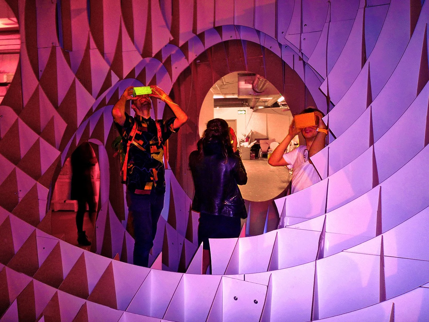 Three people standing in front of a large circular mirror with a geometric patterned frame, taking photos with VR headsets in a colorful indoor space.