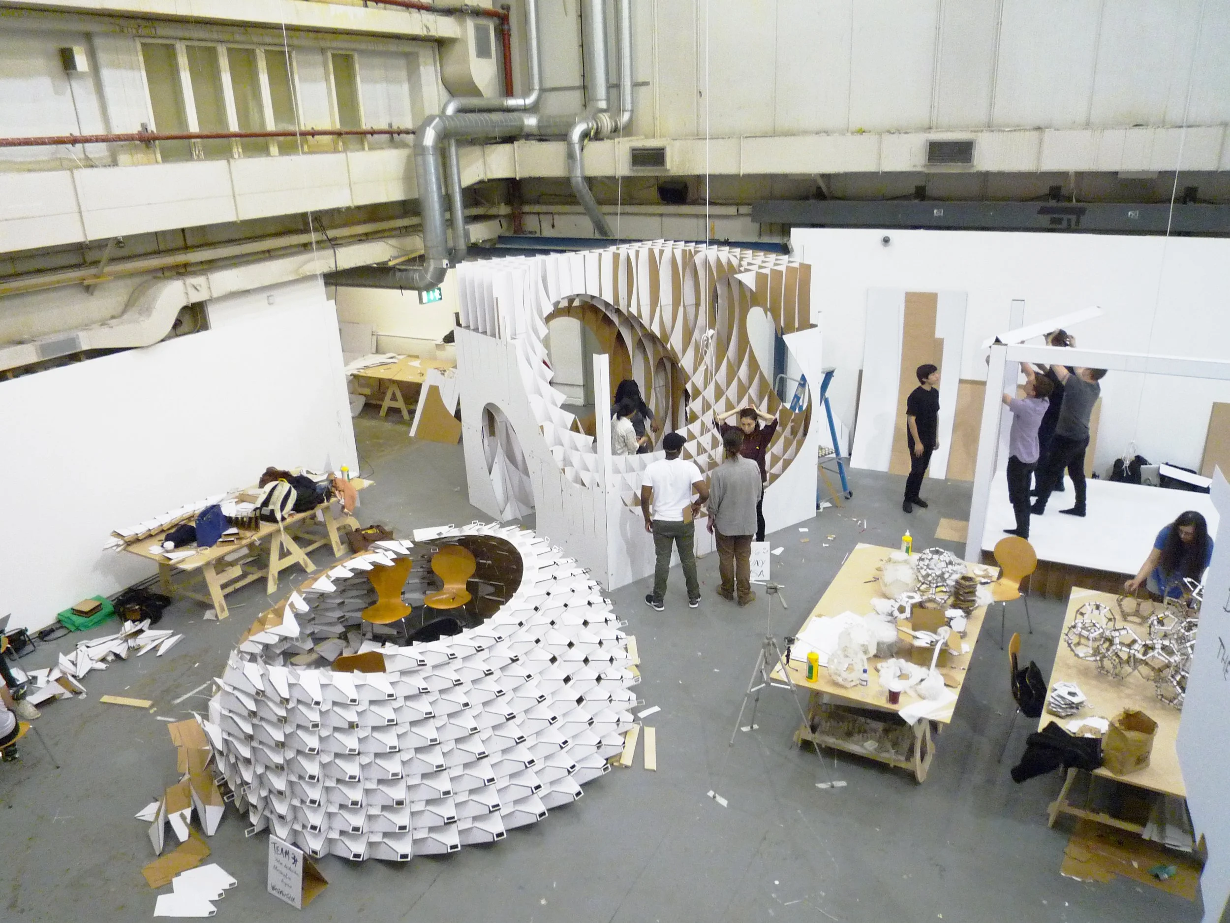 People assembling geometric wooden or cardboard arch shapes and circular structures in a large industrial workshop or warehouse with high ceilings and exposed piping.