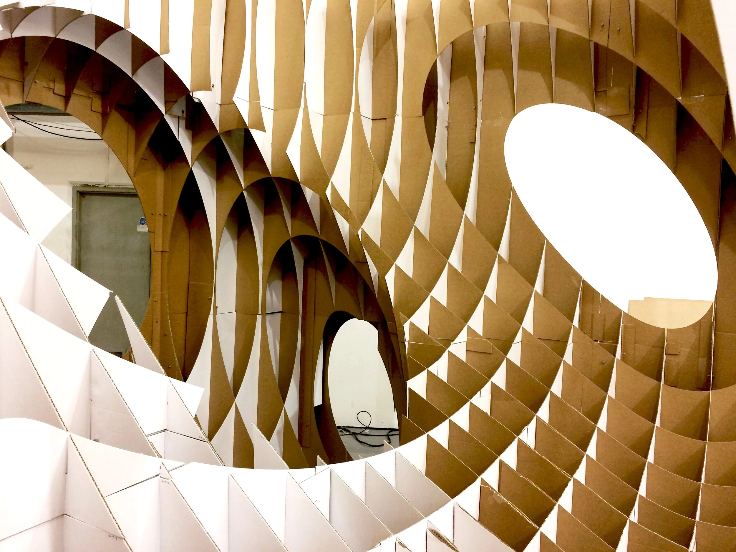 Close-up of a large, abstract geometric sculpture made of interconnected wooden panels, forming a spiraling pattern with alternating light and dark wood sections.