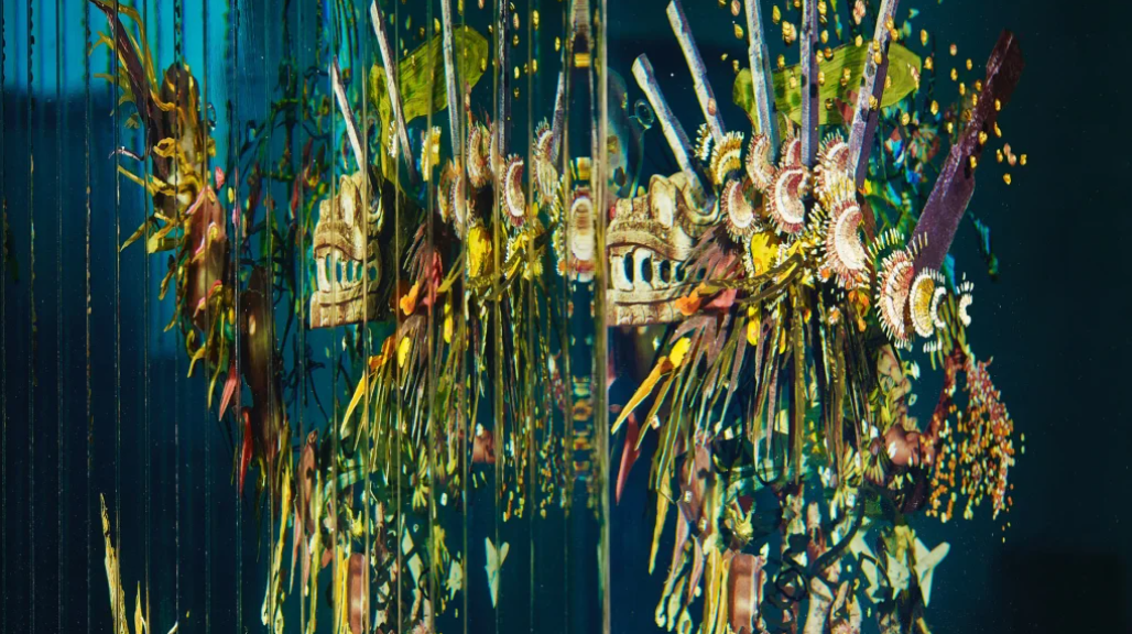 Colorful art installation with various feathers, wooden objects, and decorative elements suspended from above, set against a dark background.