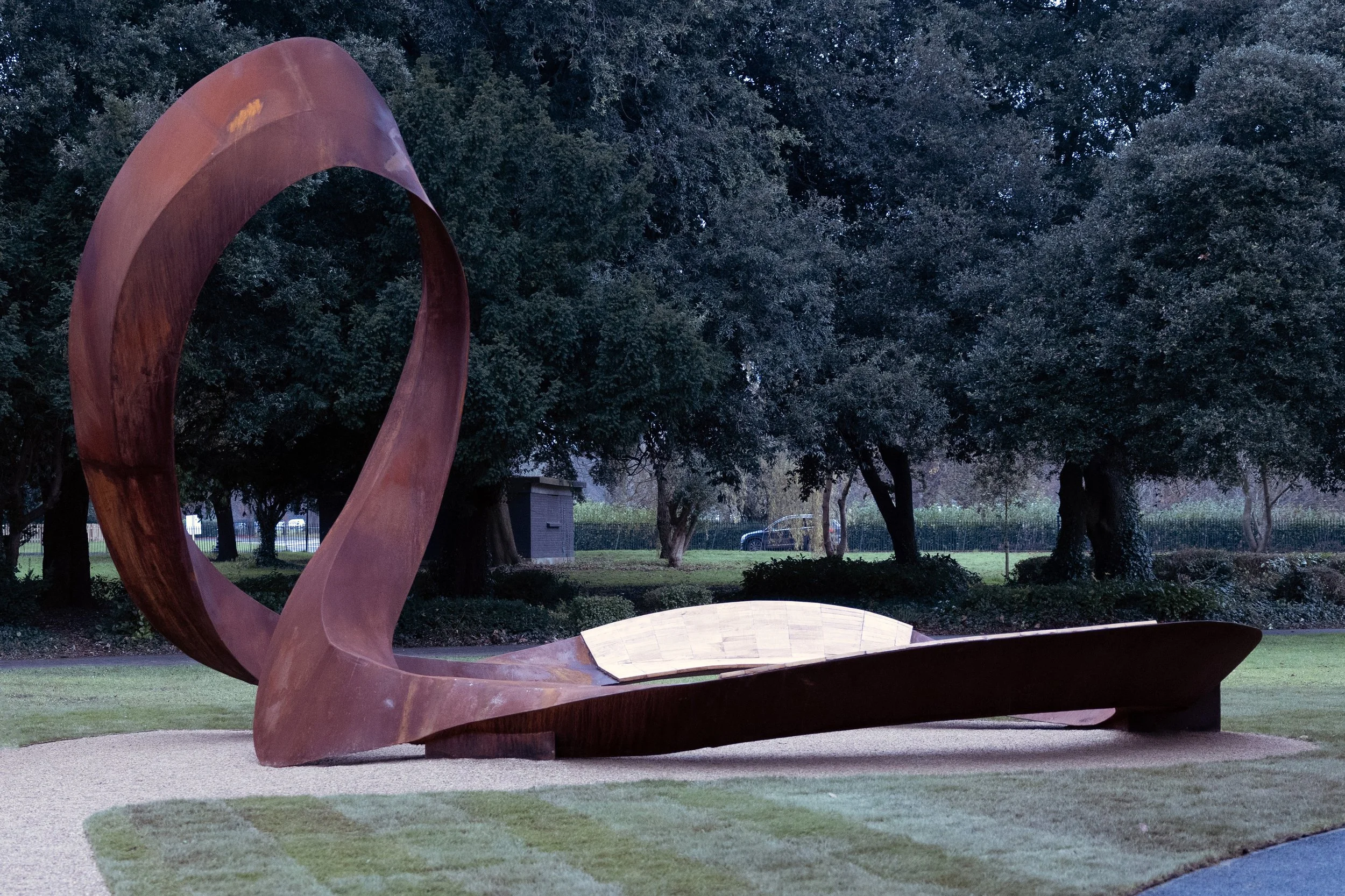 A large abstract metal sculpture with flowing, curved shapes located in a park with trees and grass, during twilight.