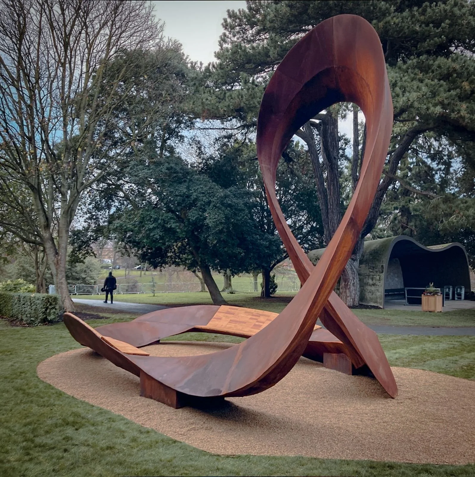 Large wooden sculpture resembling an open book with flowing pages, situated in a park with trees and grass, with a person walking in the background.