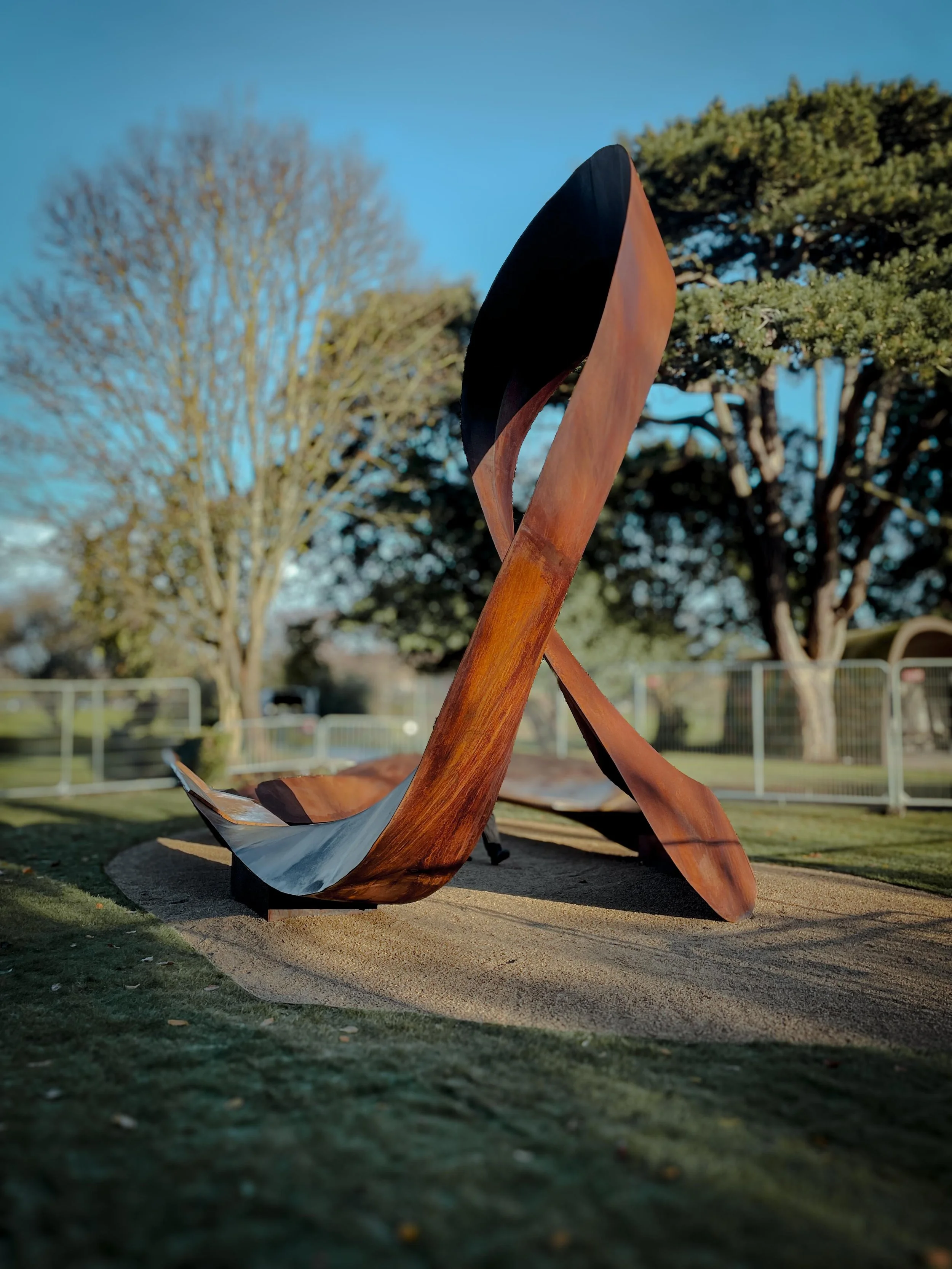 Outdoor sculpture made of wood and metal resembling a leaf or bird, set against trees and a fence on a sunny day.