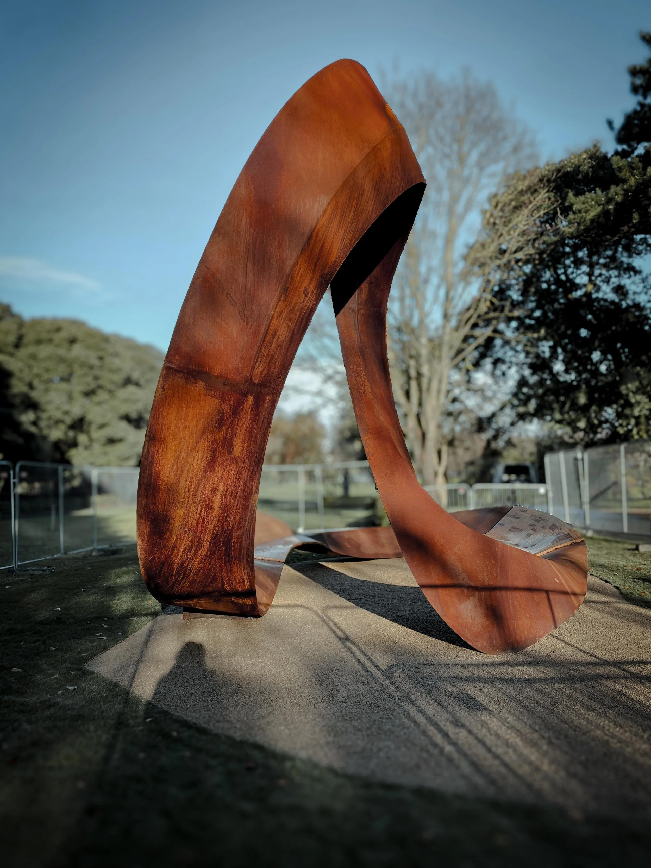 A large abstract outdoor sculpture made of rust-colored metal, with curved and flowing shapes, situated on a paved path in a park with trees in the background.