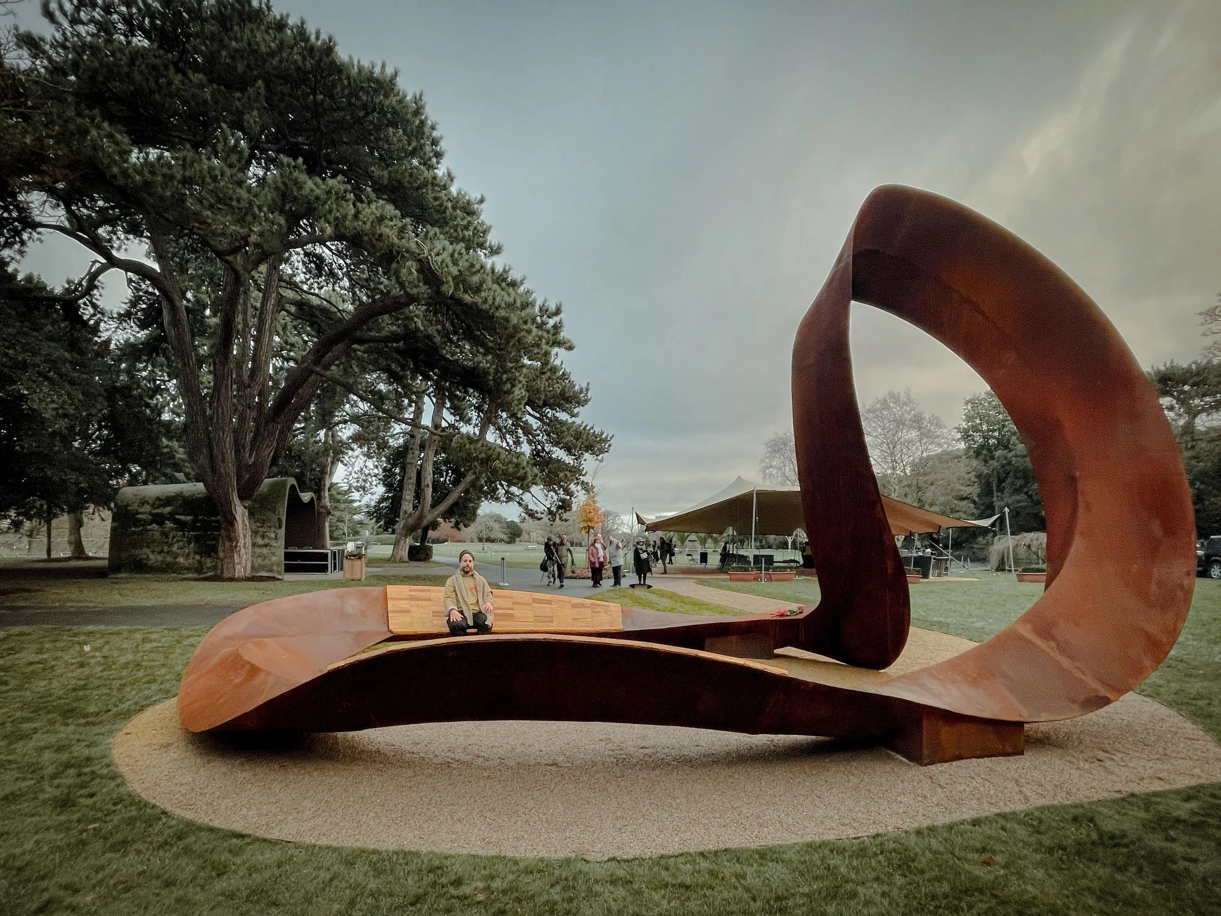 A large abstract sculpture with smooth, curved, reddish-brown surfaces is situated on a circular gravel area in a park. In the background, there are tall trees, a group of people walking, and a tent or canopy. The sky appears cloudy.