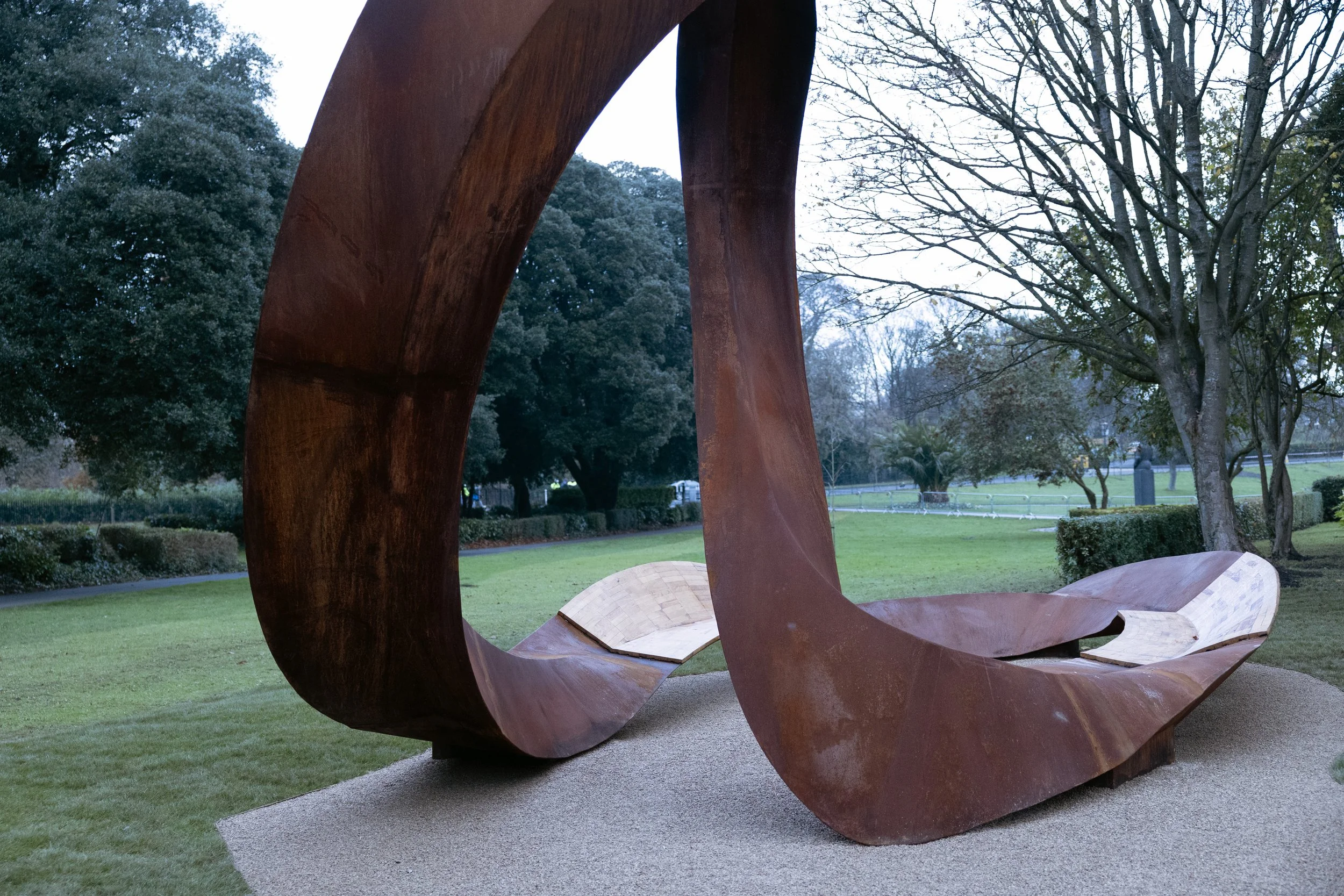 Large abstract sculpture made of rust-colored metal in a park with trees and green grass.