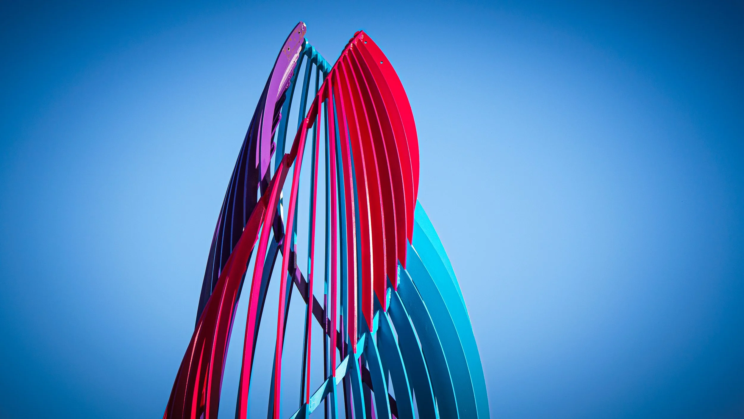 Colorful modern sculpture with elongated, curved, vertical elements in purple, red, and blue, set against a clear blue sky.
