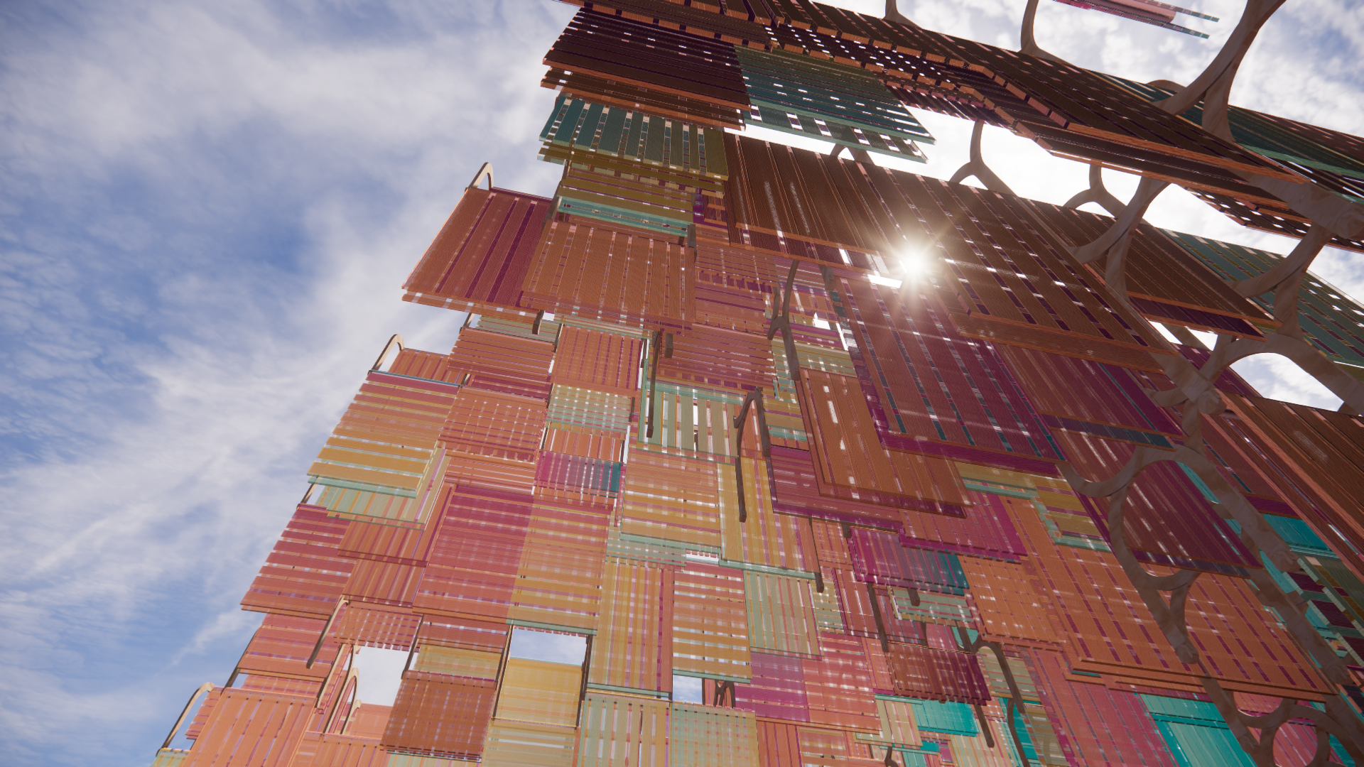 A ground-up view of a colorful art sculpture made from shipping containers arranged in a tall, abstract structure against a partly cloudy sky with the sun shining through.