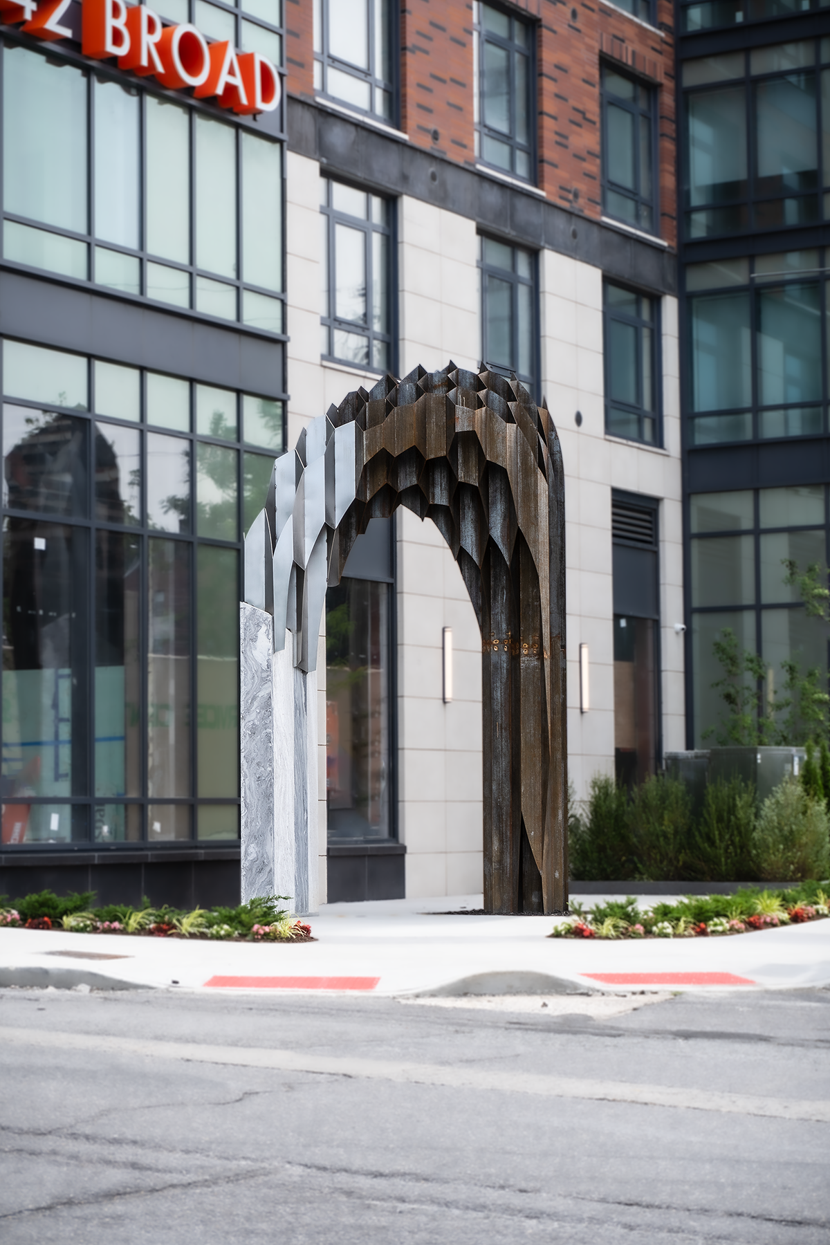A modern central city street scene featuring a decorative metal archway entrance with a honeycomb pattern, surrounded by landscaped flower beds, in front of a glass and brick high-rise building. The building has reflective windows and a red sign with