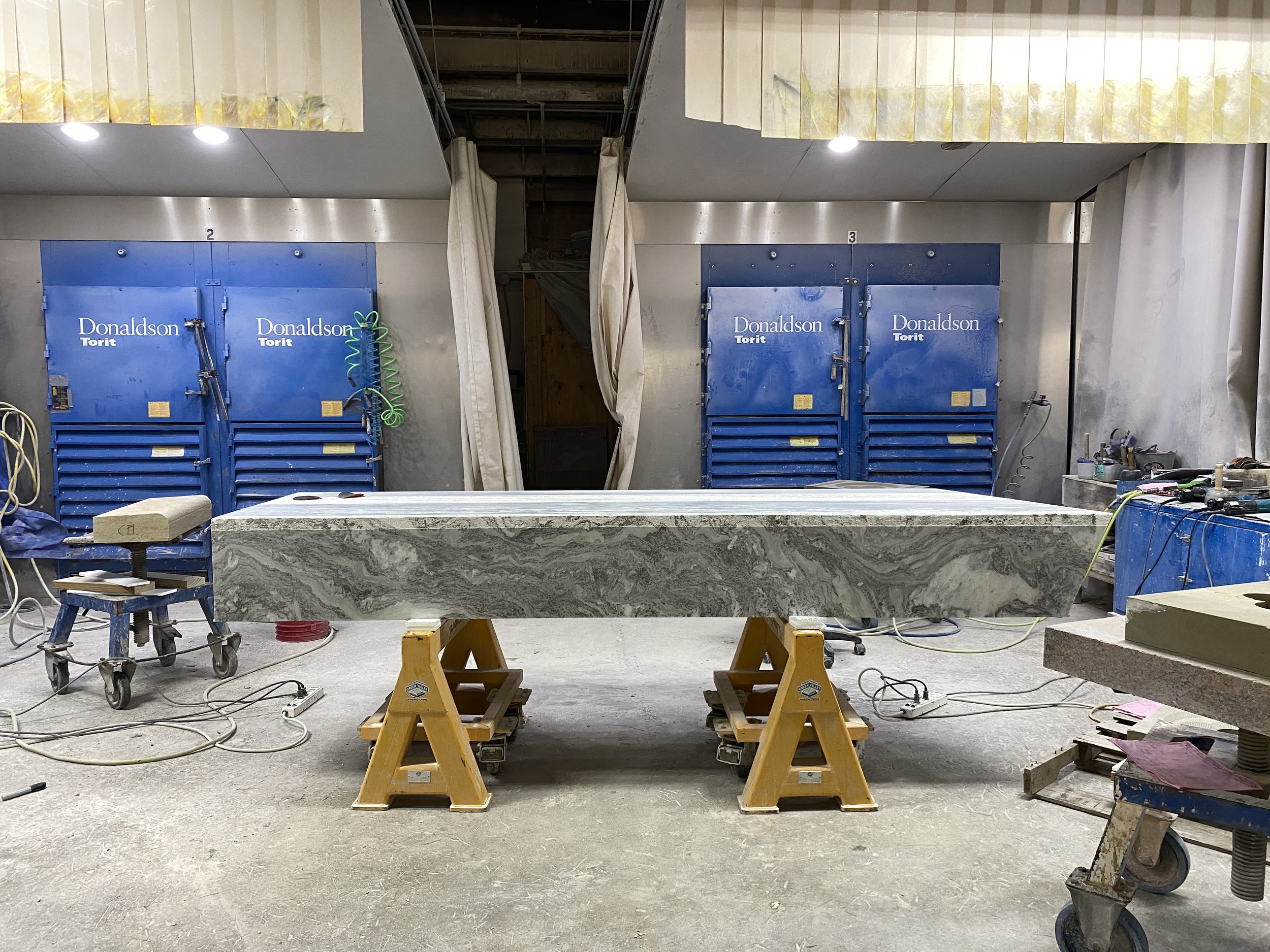 An unfinished kitchen or workspace under construction featuring blue toolbox units labeled 'Donaldson Torit.' There are stool and worktable in the foreground, with various tools and wires scattered around. Two curtains hang in the middle, covering a 