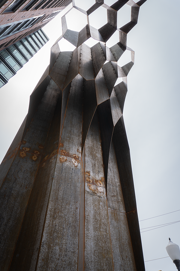 Low-angle view of a modern urban building with a rusted metal sculpture resembling a tree trunk with hexagonal shapes on top.