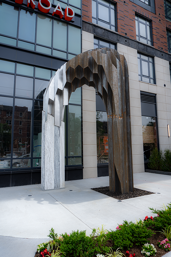 Modern outdoor sculpture of an arch made of metal and stone, located in front of a contemporary building with large glass windows and brickwork.